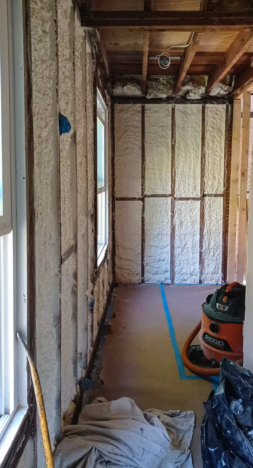 A room under renovation with framed walls and foam insulation. Two windows on the left, a blue tape line on the floor, and a shop vacuum cleaner in the corner.