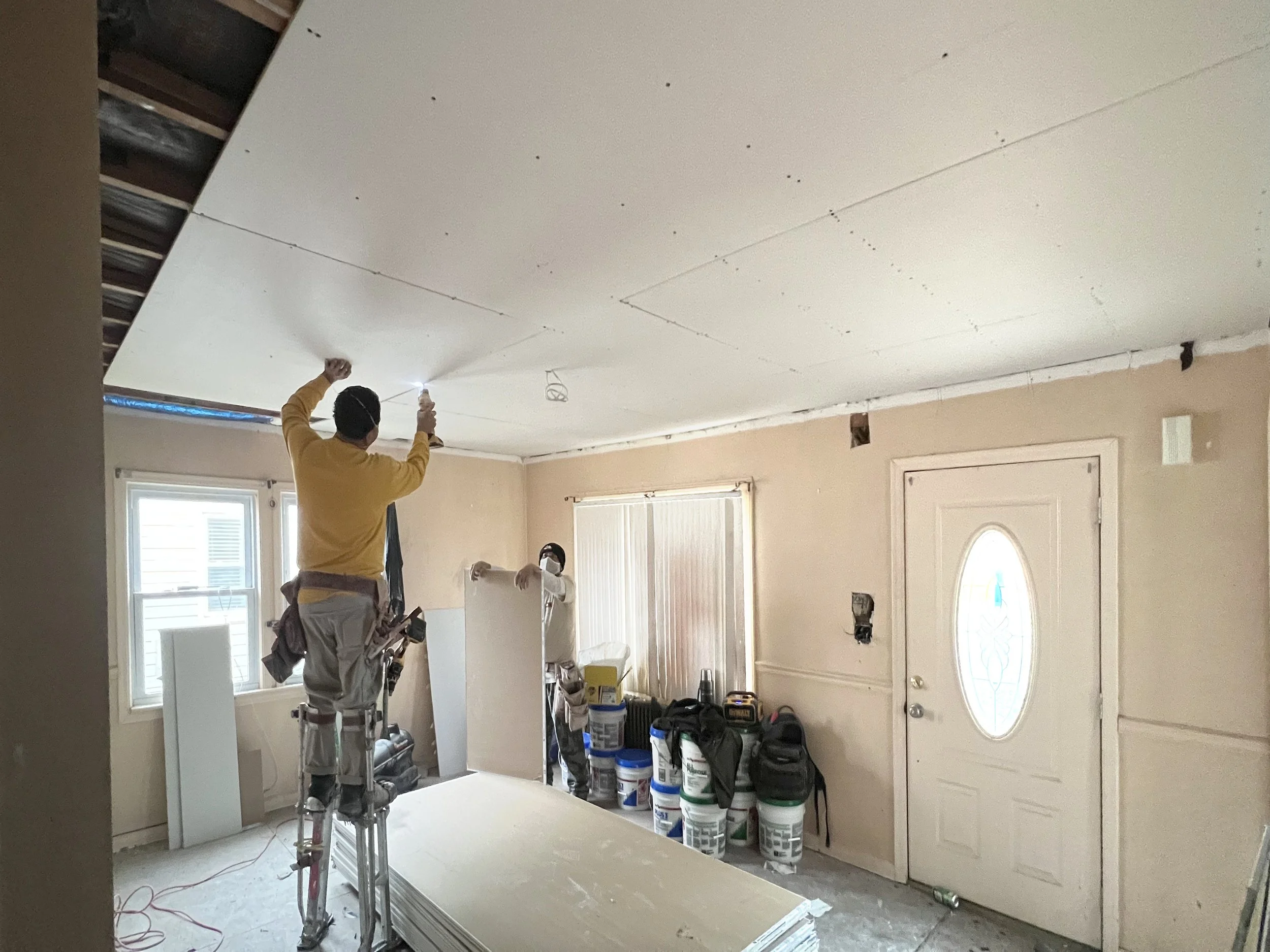 Two workers installing drywall in a room with beige walls, a white door, and two windows. One worker stands on a step ladder, using a tool on the ceiling, while the other leans on a drywall panel. The room contains construction supplies and tools.