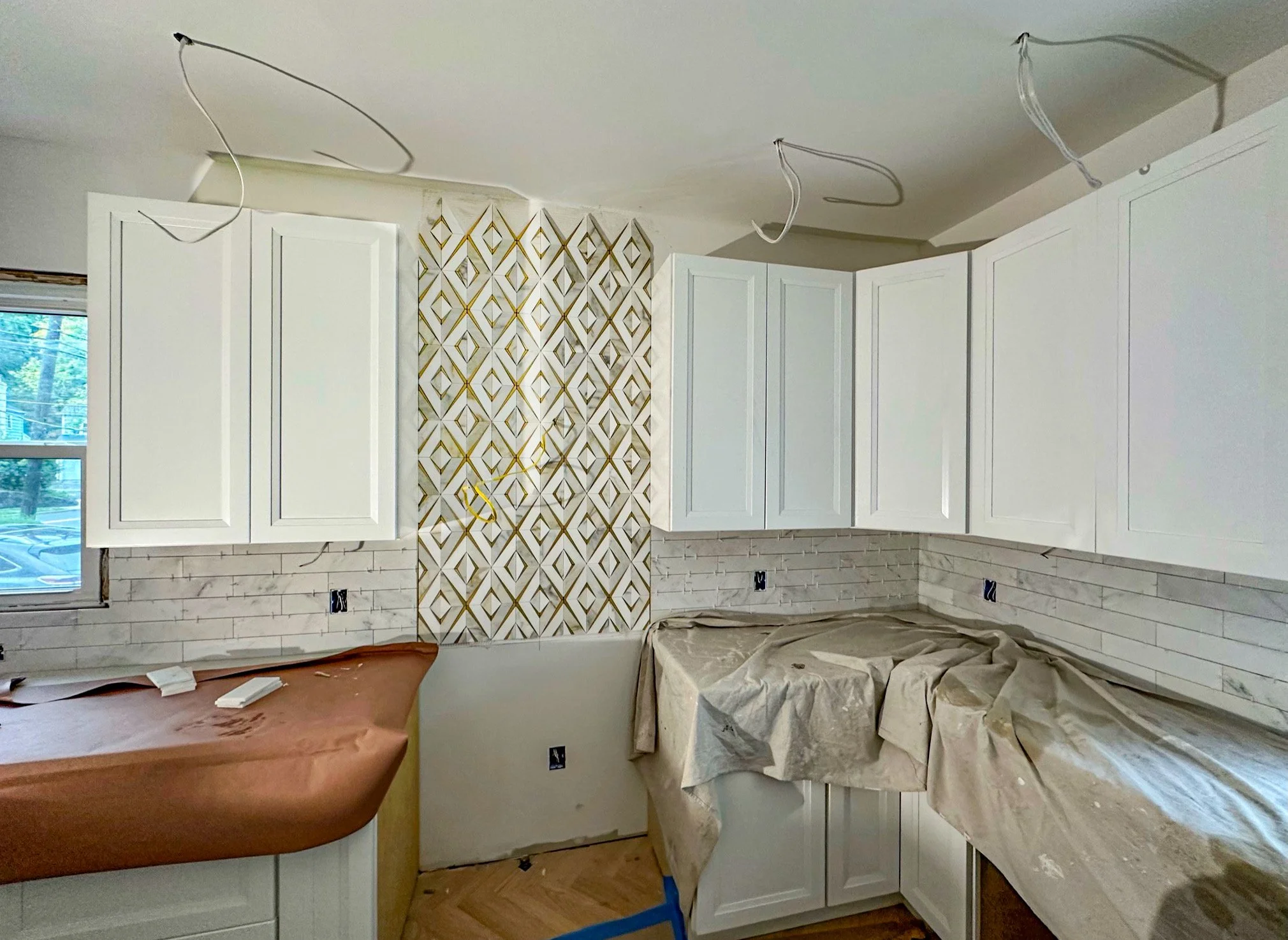 Kitchen under renovation with white cabinets, a patterned tile accent wall, and covered countertops, wires hanging from the ceiling.