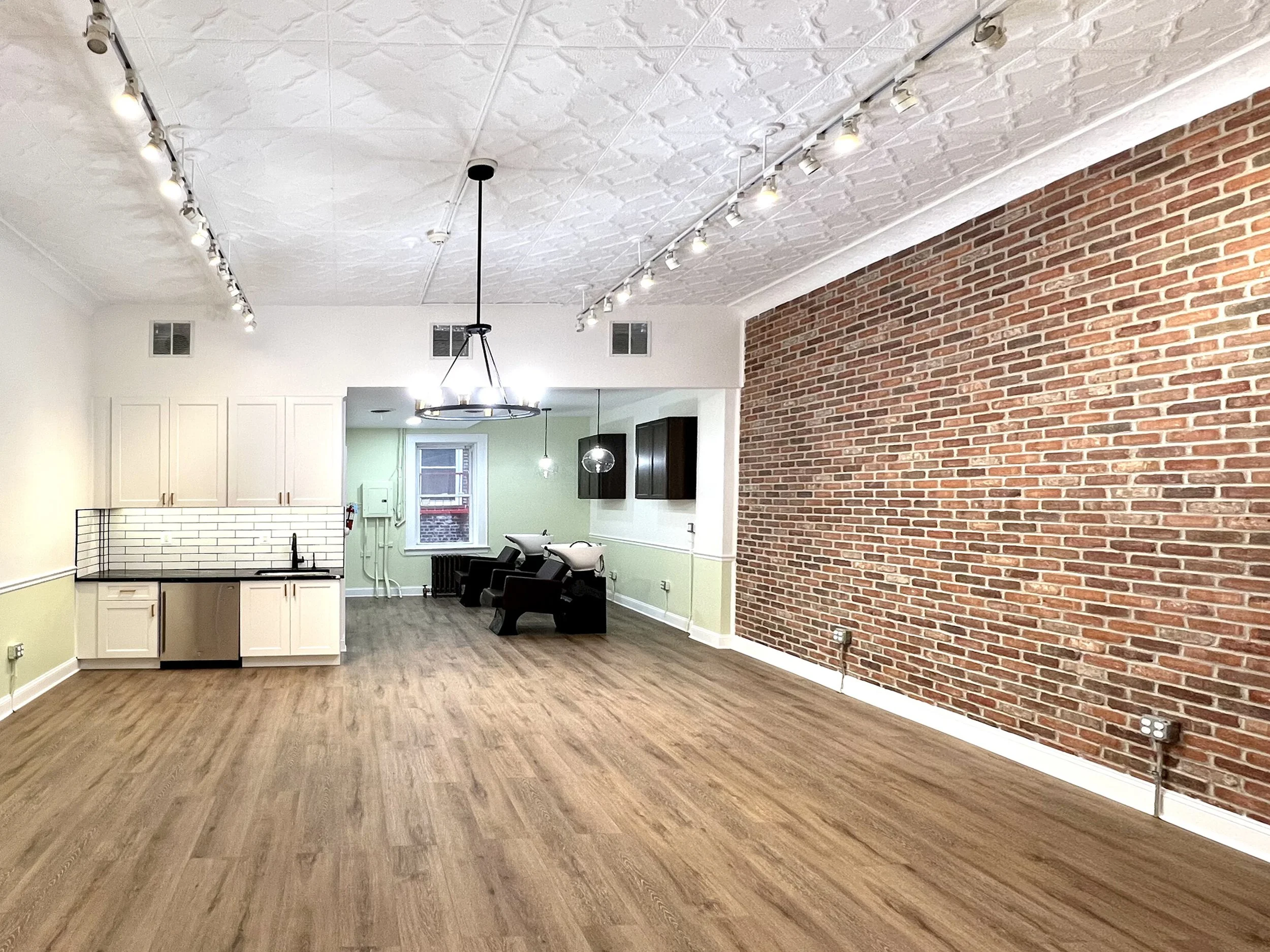 Empty room with wooden floors, a brick accent wall, a kitchenette with white cabinets, black countertops, and a black sink, track lighting on the ceiling, and two black salon chairs with wash basins in the background.