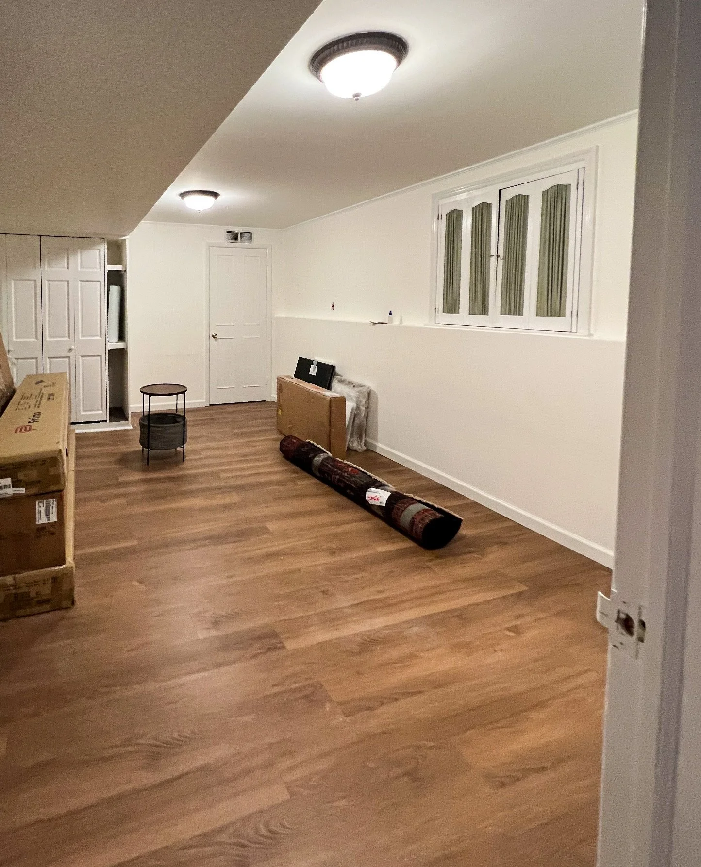 Empty room with hardwood floors, a closed door, a small table, boxes, and rolled rug, with a window and ceiling lights.