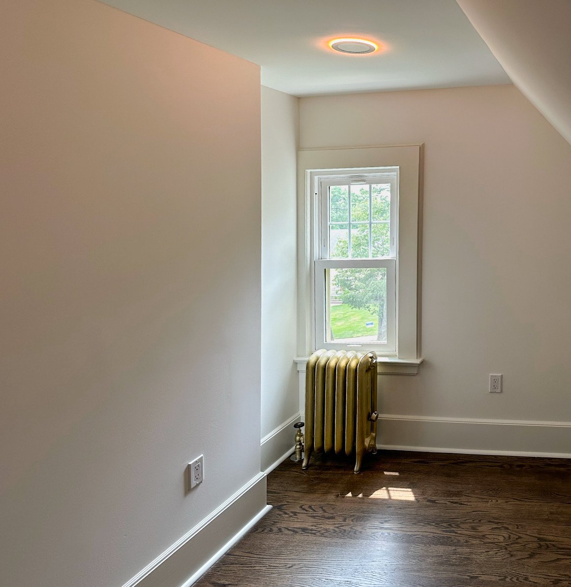 Empty room with hardwood floor, white walls, a single window letting in natural light, and an old-style yellow radiator near the window.