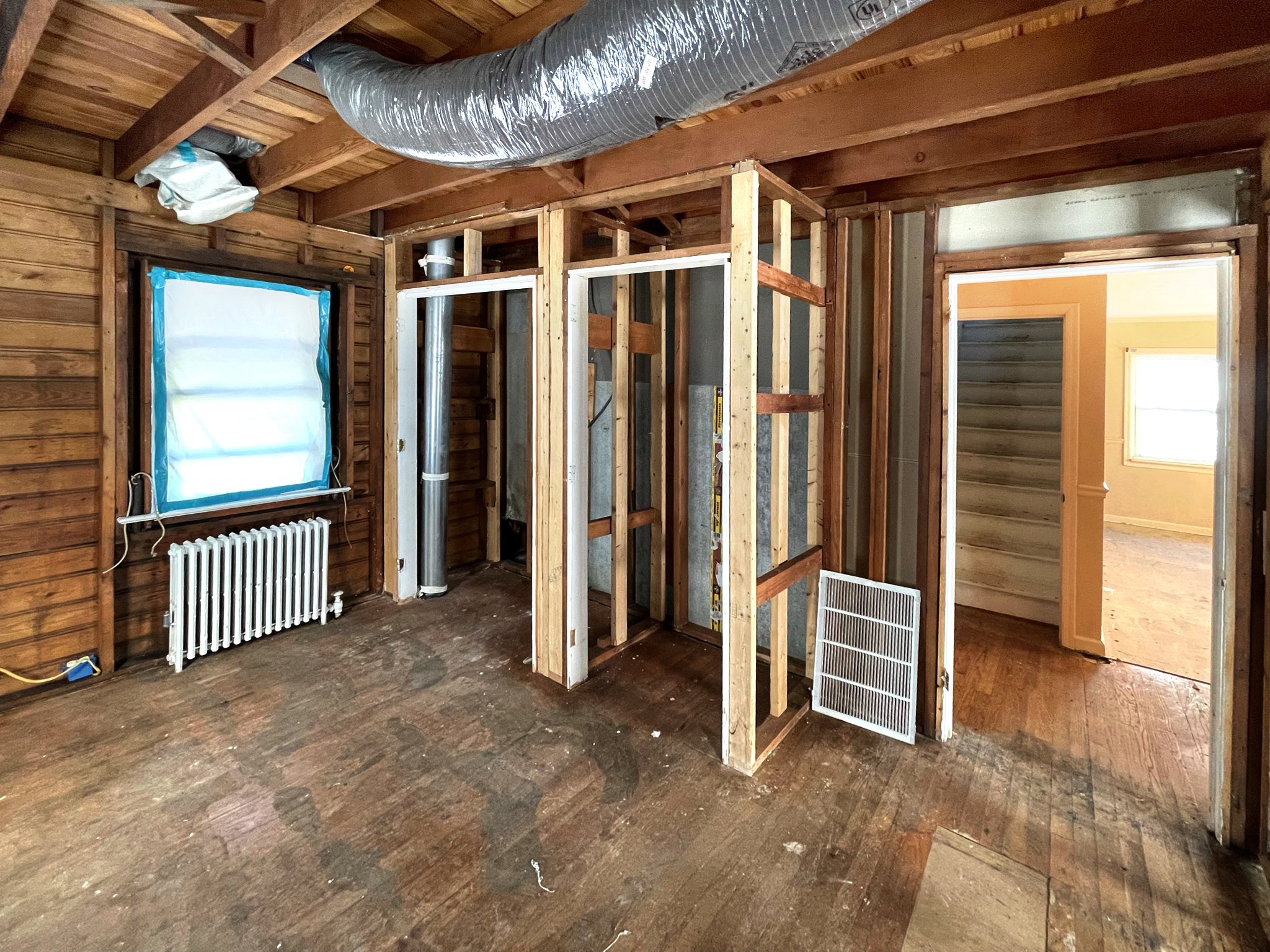 Room under renovation with exposed framework, wooden walls, a window with blue plastic covering, and a doorway leading to another room with a window, hardwood floors, and wall panels.