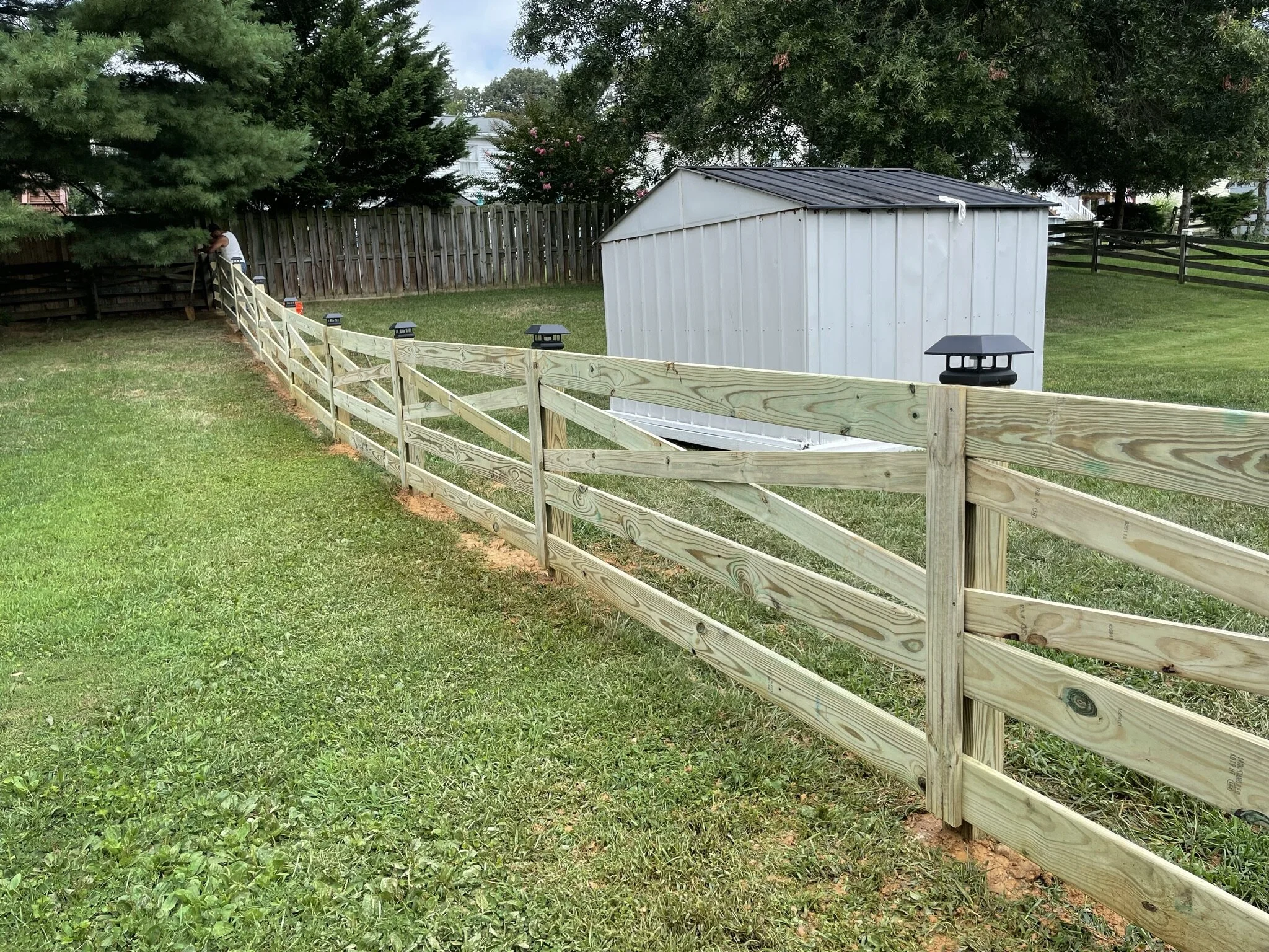 A backyard with a newly built wooden fence, a small white metal shed, trees, and a person working near the fence.
