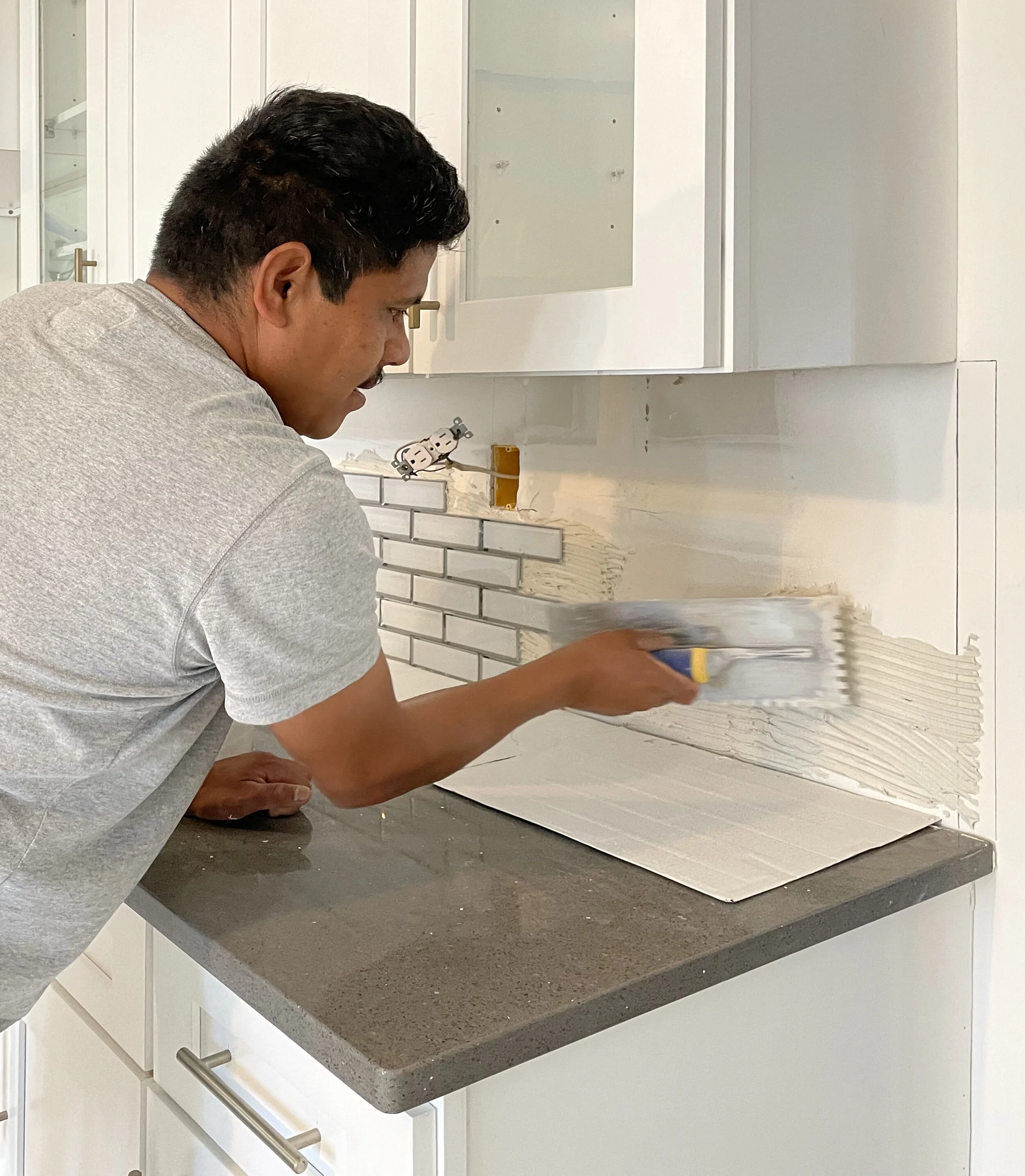 A man in a gray t-shirt applying white tile adhesive to a wall with a notched trowel for installing backsplash tiles in a kitchen.