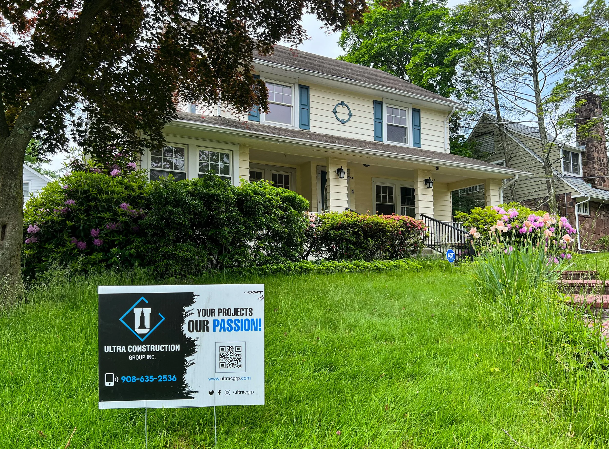 A yellow two-story house with blue shutters and a front porch, surrounded by green bushes and colorful flowers. A sign in the yard displays construction company information.