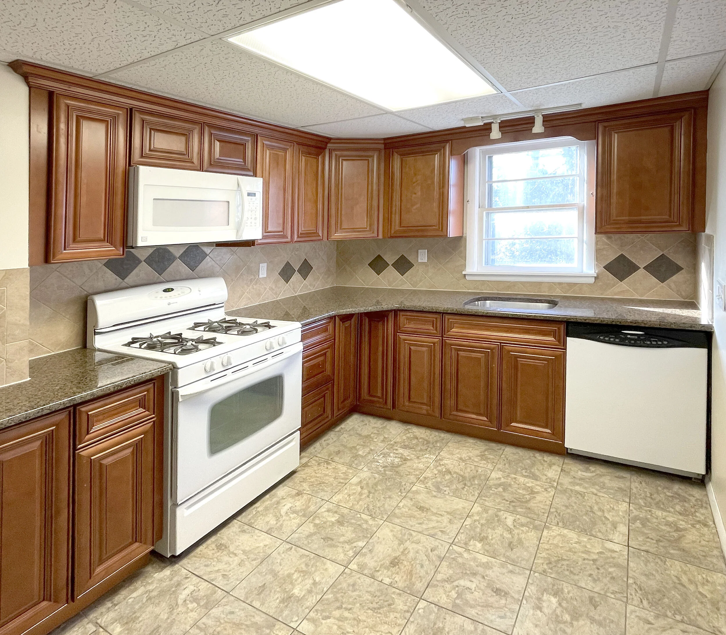 Kitchen with brown wooden cabinets, beige tile floor, beige tiled backsplash with diamond accents, white stove and microwave, and a window above the sink.