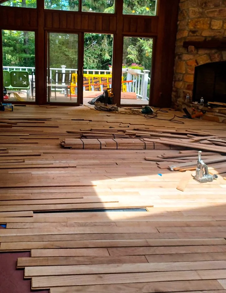 Inside a house with large windows and a stone fireplace, a wooden floor is being installed as part of a renovation project. Tools and wood planks are scattered across the floor.