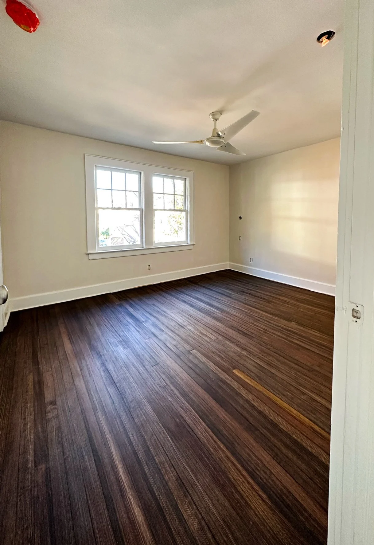 Empty room with dark wood floor, white walls, two large windows, a ceiling fan, and a partially visible door.
