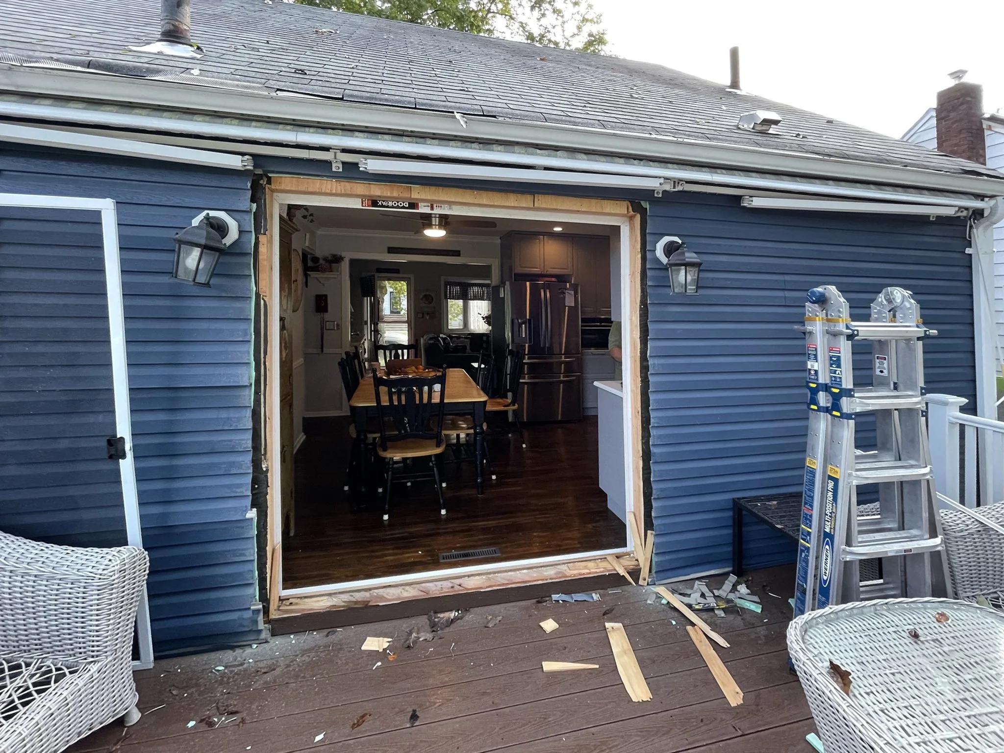 Back of house undergoing renovation with open door frame, construction debris on deck, ladder, and outdoor furniture.