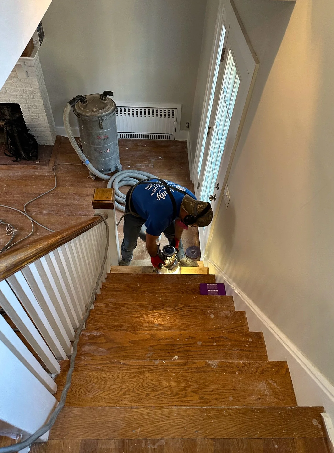A person is sanding a wooden staircase while using a handheld sander. The staircase has a wood finish, and there are dust and debris on the steps. The person is wearing gloves and ear protection, with a large vacuum hose connected to a vacuum device 