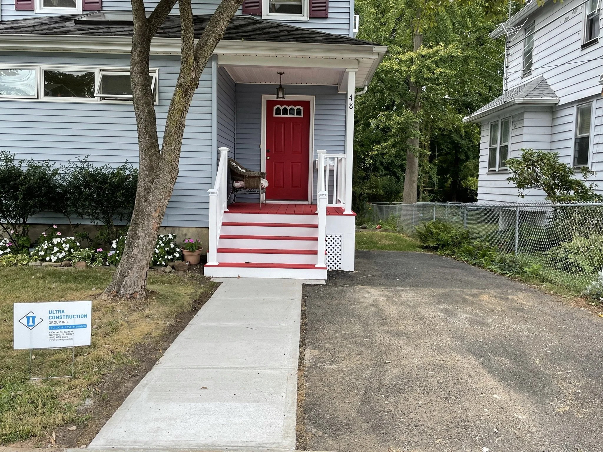 Front of a house with gray siding, red front door, and a small porch. A sign in the lawn advertises Ultra Construction. A tree partially obscures the porch, and there are flowers along the lawn.