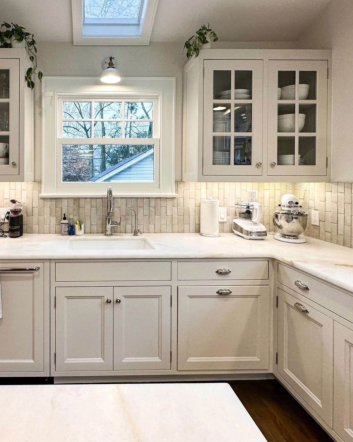 White kitchen with upper cabinets with glass doors, a window above the sink, small potted plants on upper cabinets, and kitchen appliances including a coffee maker, stand mixer, and a food processor on the countertop.