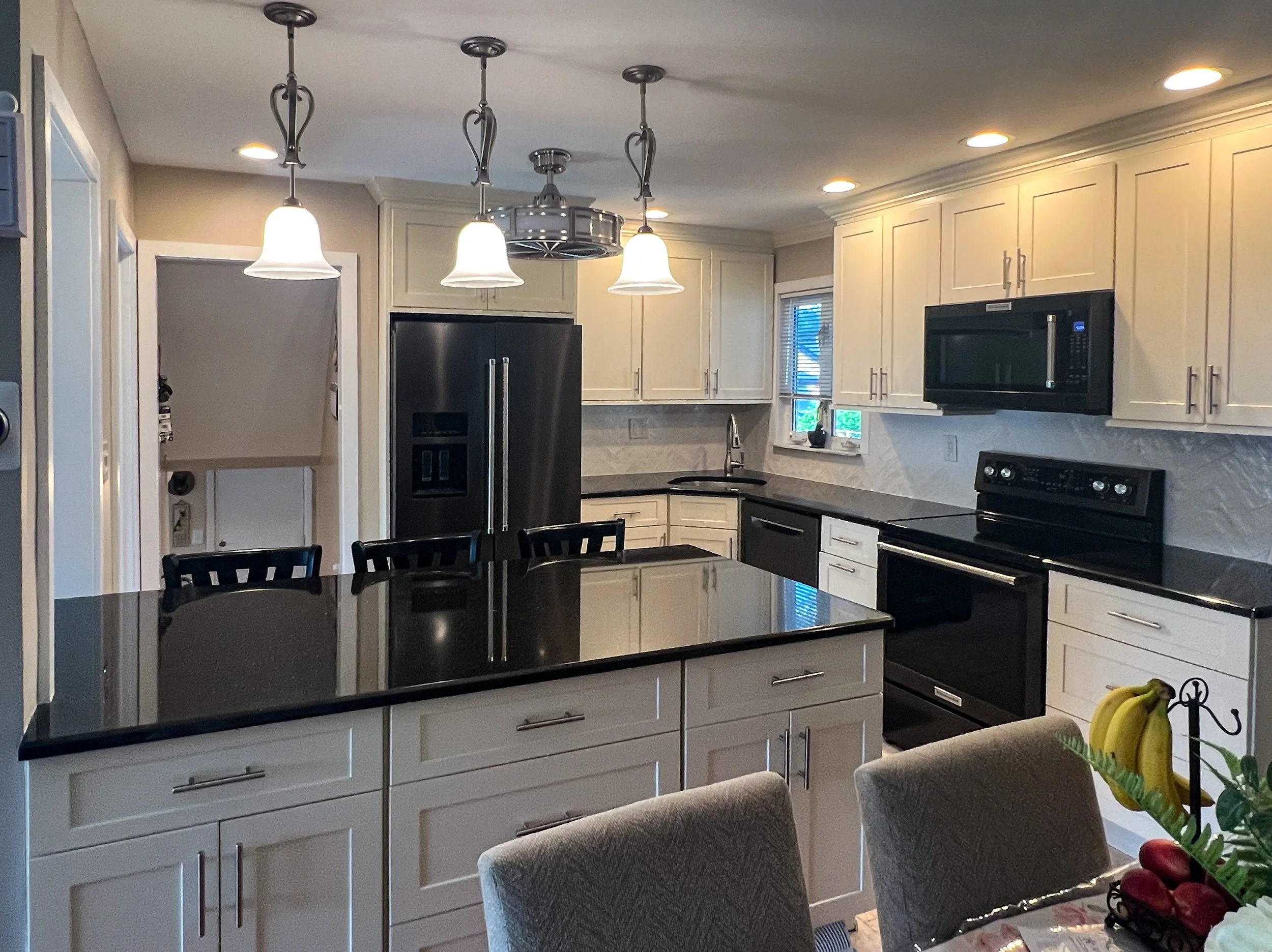 Kitchen with white cabinets, black countertops, and black appliances including a refrigerator, microwave, and oven. There is a kitchen island with a black countertop, three chairs, and a hanging light fixture with three bulbs.