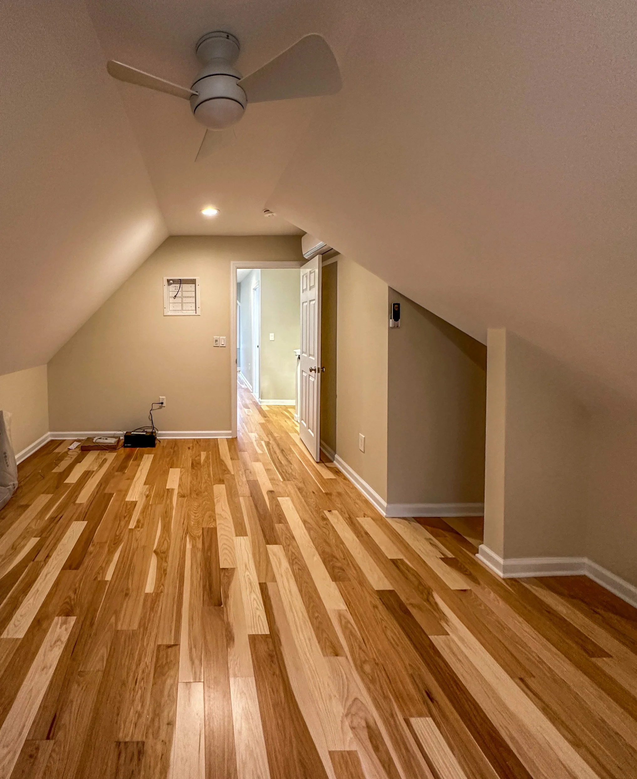 Empty attic room with wood flooring, sloped ceiling, ceiling fan, open door leading to a hallway, wall-mounted remote, and a small wall panel.