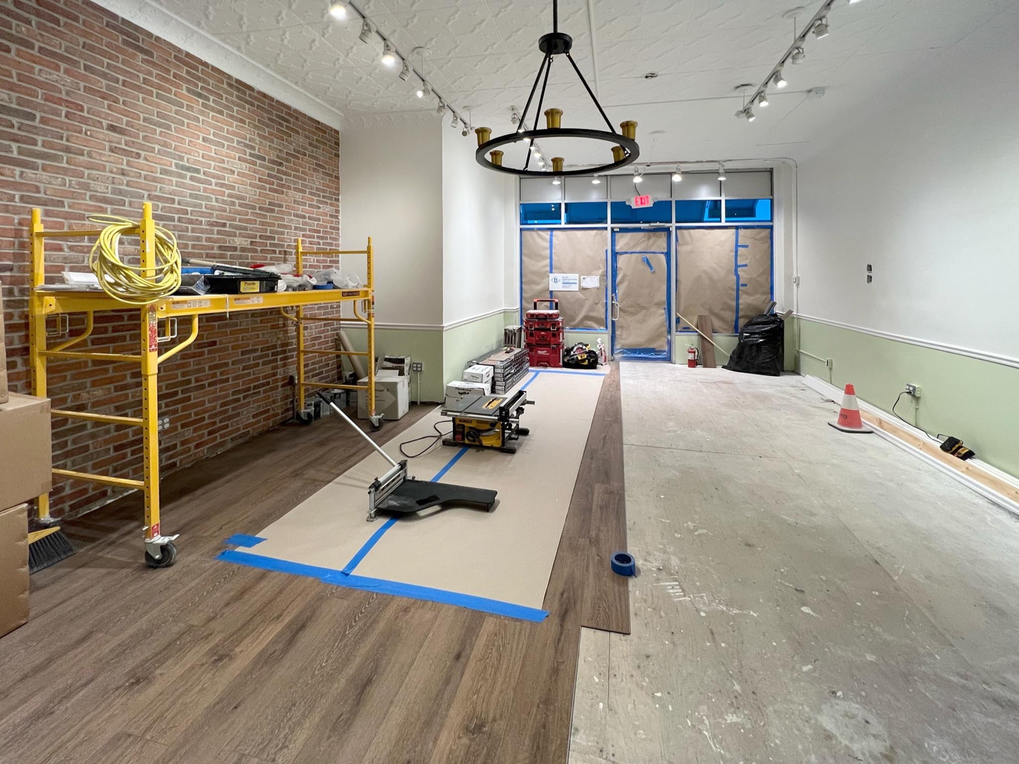 Interior room under renovation with partially installed wood flooring, construction tools, a yellow scaffolding, and construction supplies. The room has a brick wall on the left, a white wall on the right, and a large window and door covered with bro