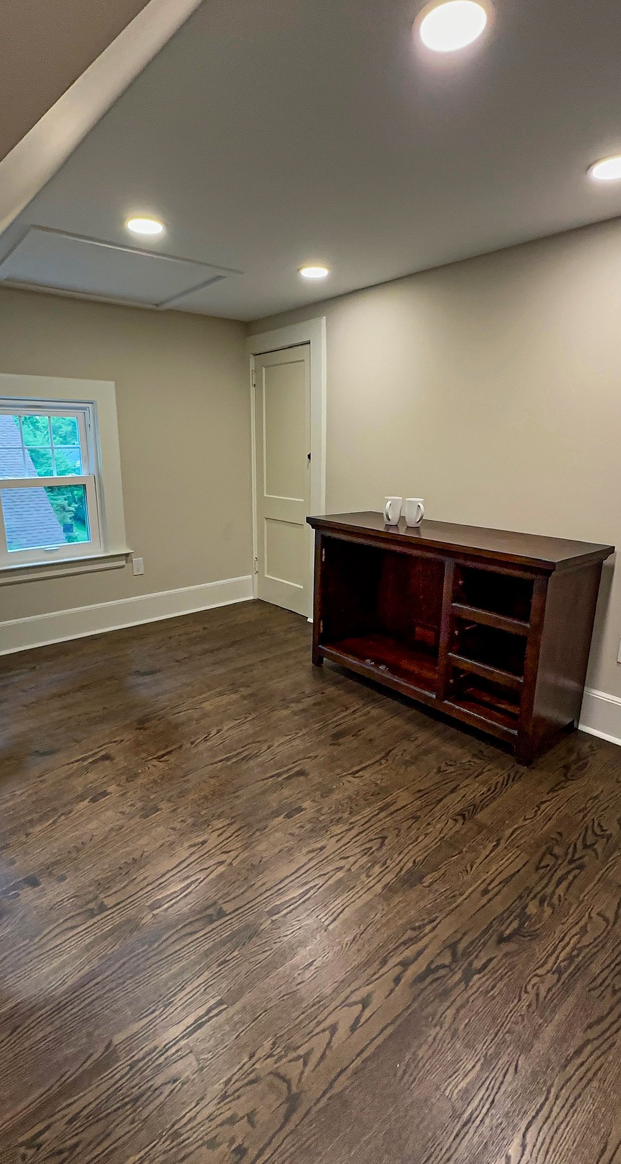 Empty room with hardwood floors, beige walls, a window, a closed white door, and a dark wooden cabinet.