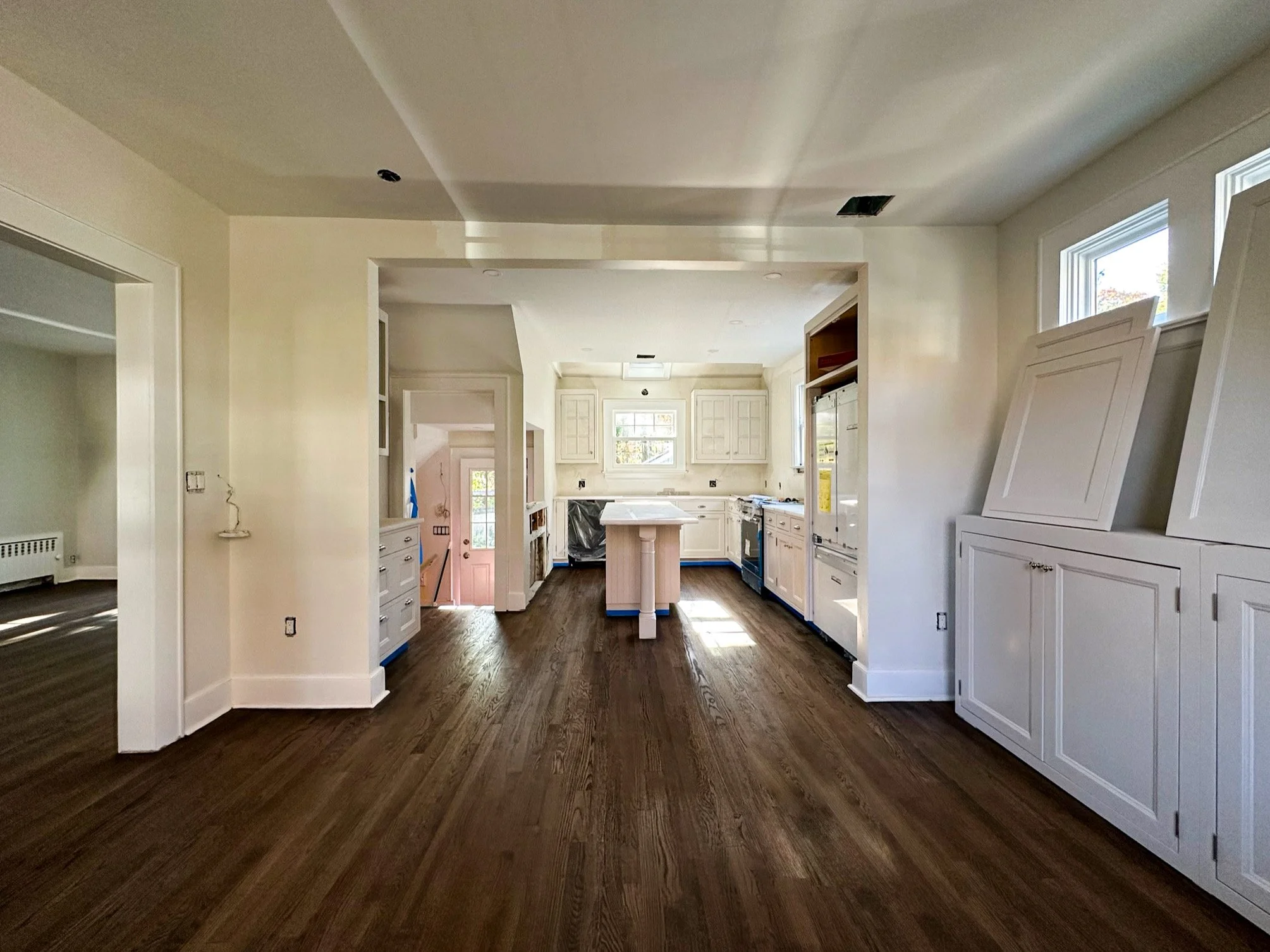 Empty kitchen with white cabinets, dark hardwood floors, and a small island in the center. Light coming through the windows, some cabinetry is still open or missing doors.