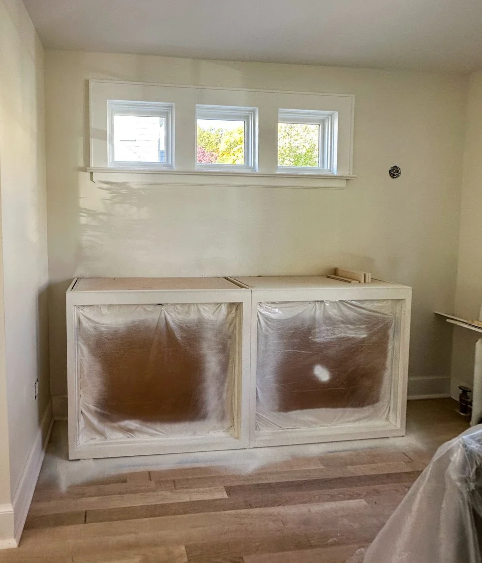 Kitchen or laundry room renovation in progress, with white cabinets covered in protective plastic, and three small windows above, allowing natural light.