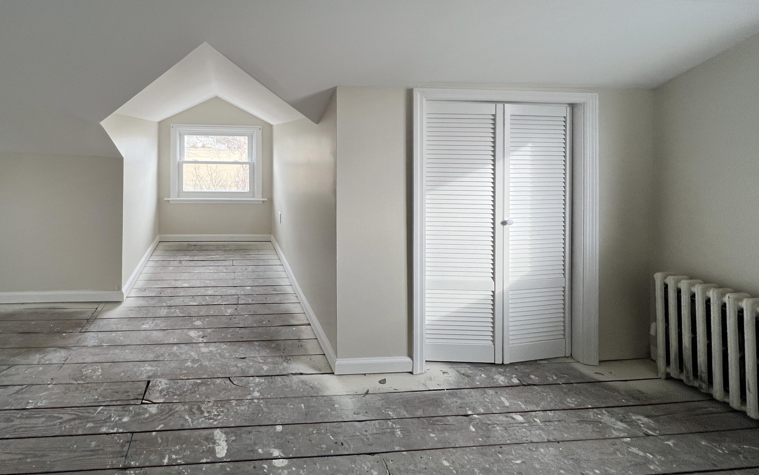 Empty, unfinished room with wooden floor planks, a small window, white walls, a closet with louvered doors, and a radiator on the right.