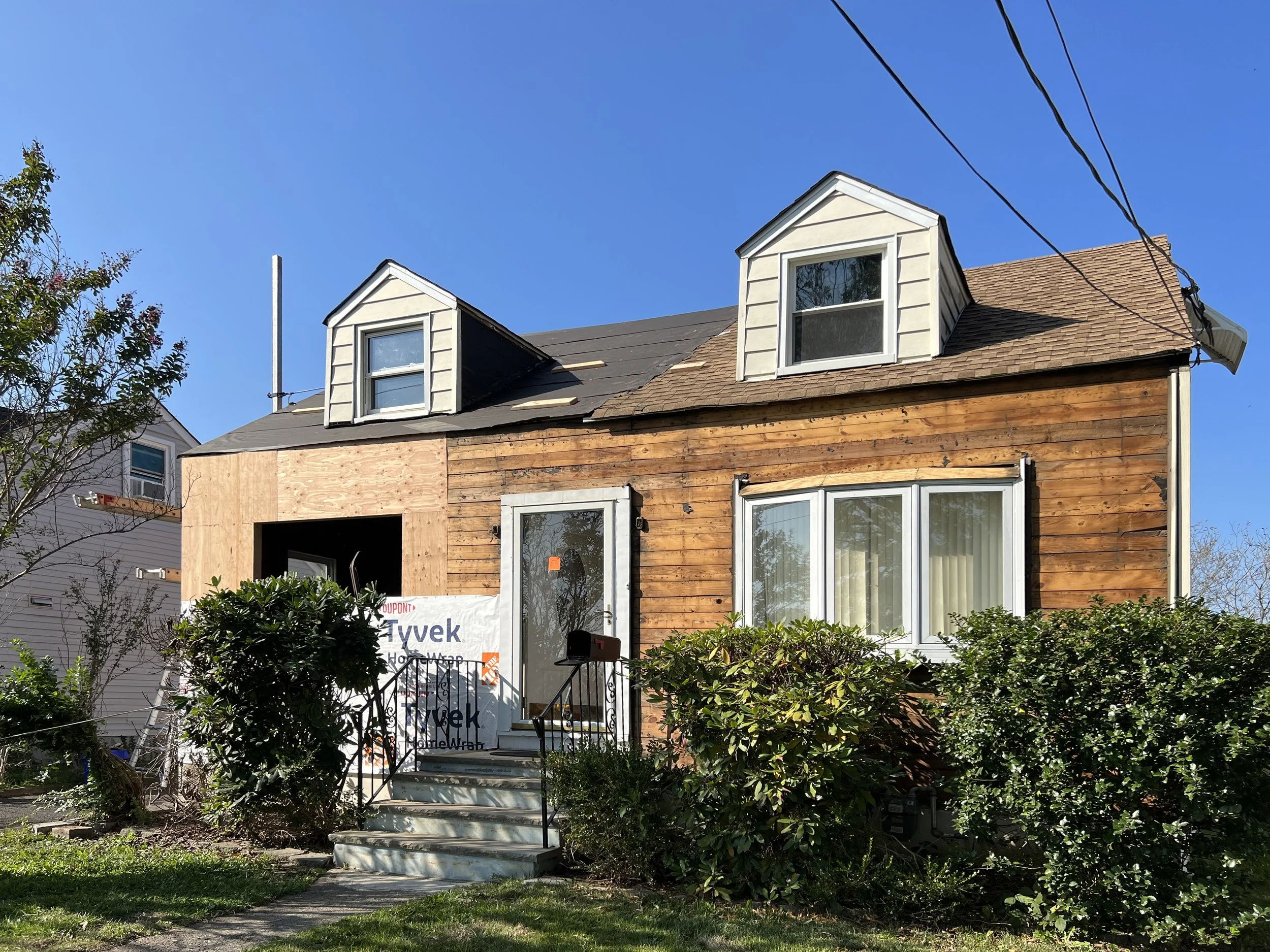 Two-story house under construction with a partial wooden and siding exterior, front steps, and a large front window, surrounded by shrubs and trees, with a clear blue sky above.
