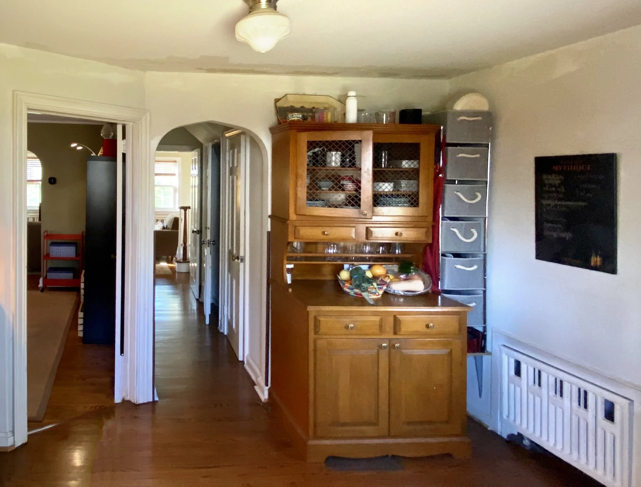 A kitchen corner with a wooden cabinet holding glassware, a bowl of fruit on top, and a storage unit beside it. To the right, there's a blackboard on the wall. The room has wood flooring and white walls.