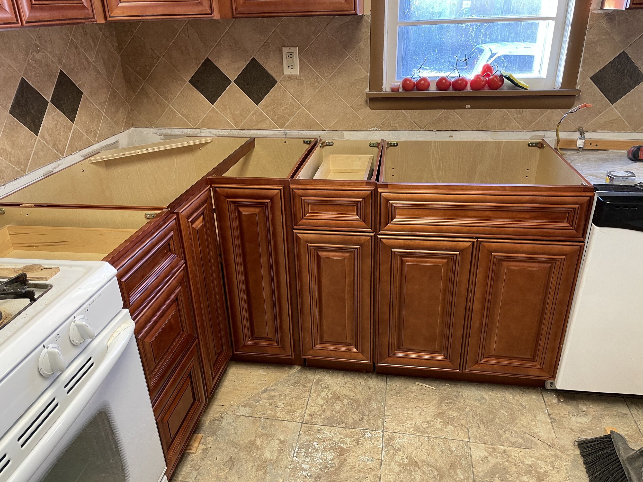 Kitchen with wooden cabinets, some without countertops, a window with tomatoes on the windowsill, and partially installed kitchen fixtures.