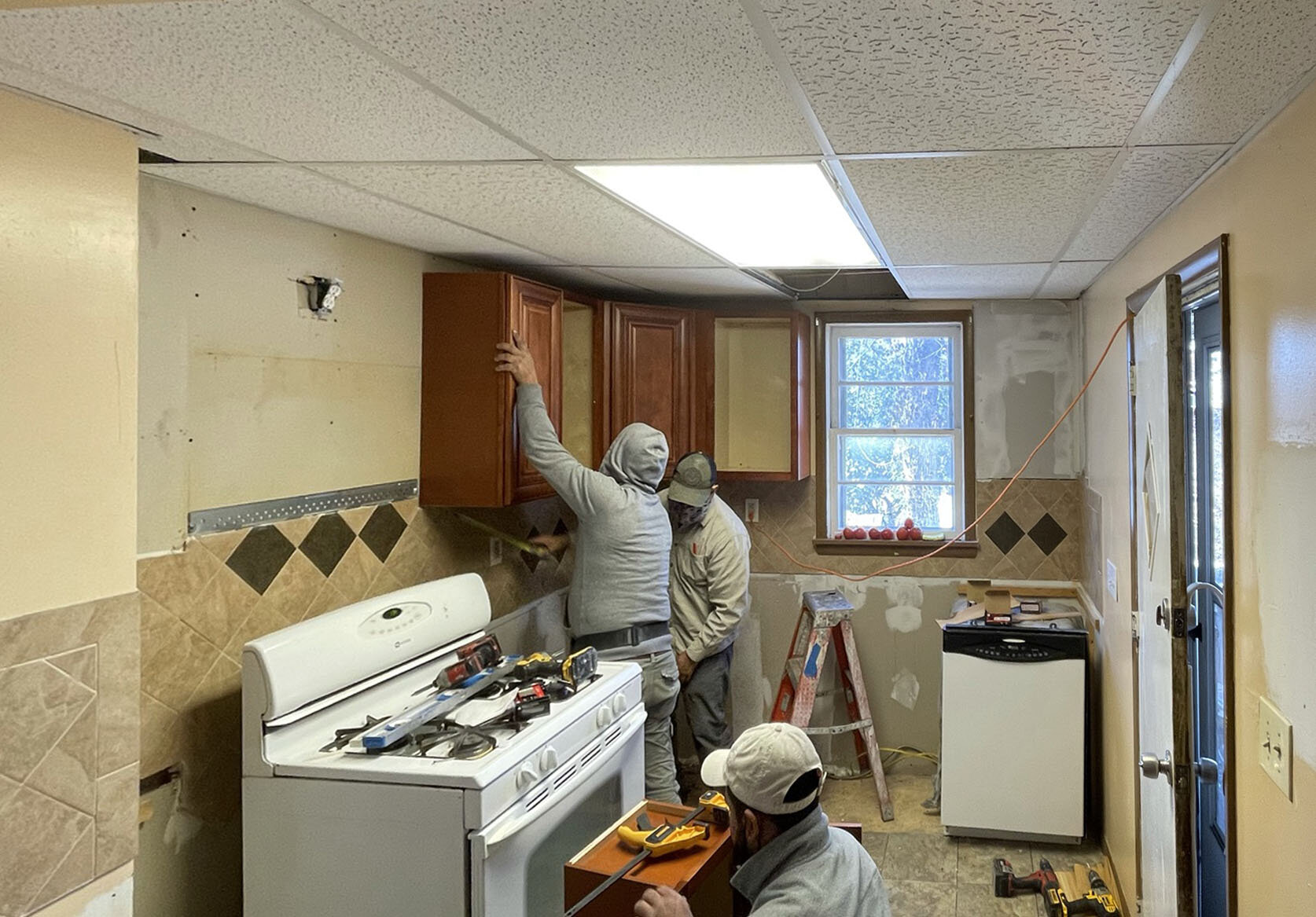Two workers installing kitchen cabinets in a partially renovated kitchen, with tools on the stove and a person in the foreground working.
