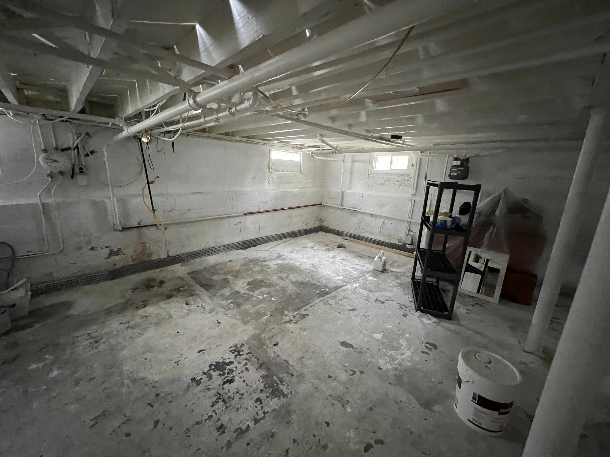 Empty basement with unfinished concrete floor and walls, exposed pipes and wires, small windows, and a black shelving unit with tools and supplies.