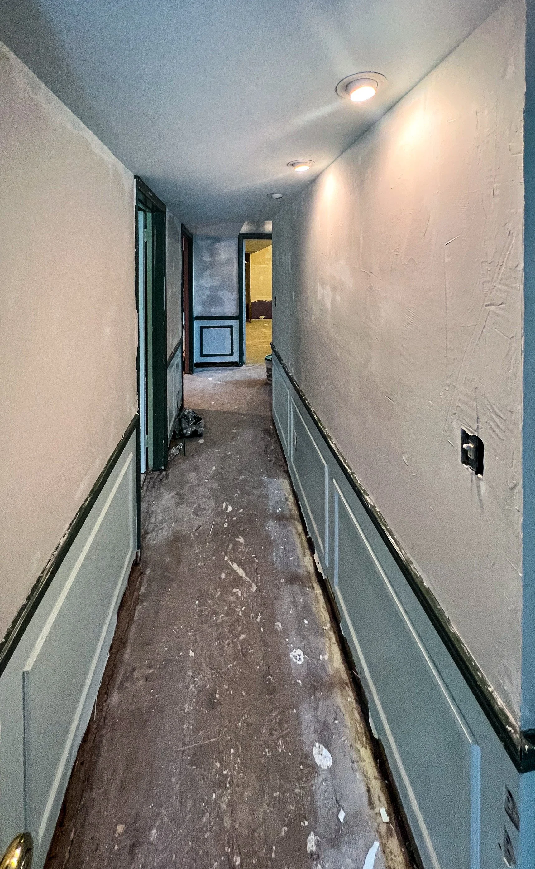 Hallway under renovation with partially painted lower wall panels, unfinished floor, and drywall patches on the walls, illuminated by recessed ceiling lights.