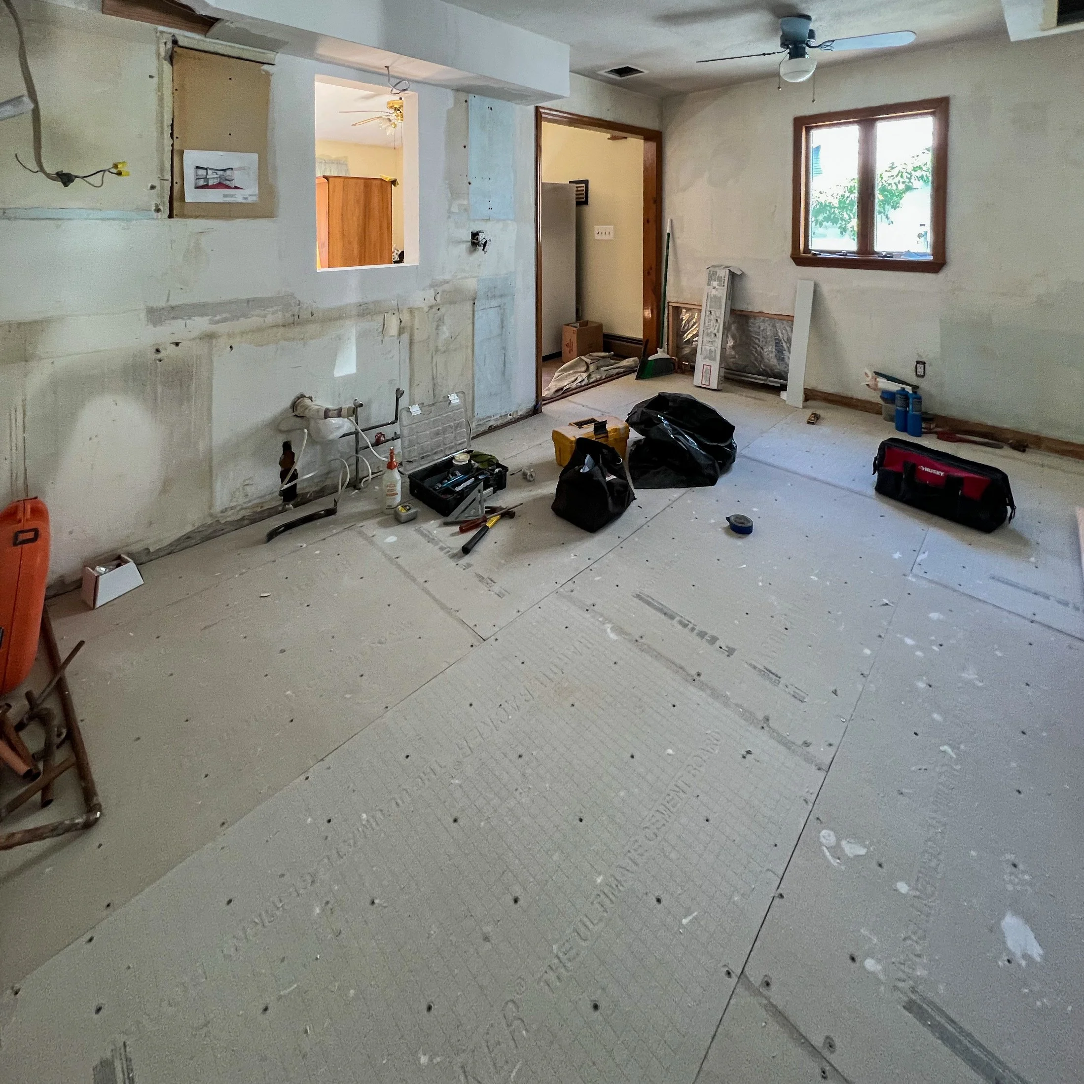 Room under renovation with exposed drywall, construction tools, and supplies scattered on the floor, including toolboxes, bags, and paint cans.