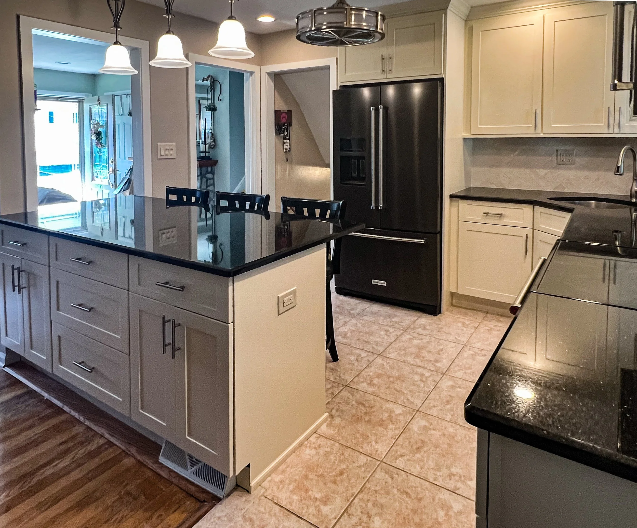 Modern kitchen with black granite countertops, white cabinets, stainless steel refrigerator, and black appliances. There is a kitchen island with white cabinetry and black countertop, three black chairs, and three pendant lights hanging above. The fl