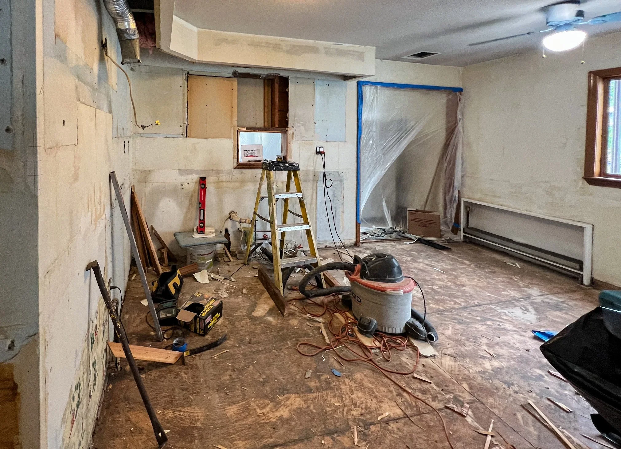 Room under renovation with construction tools, equipment, and debris on the floor, partially stripped walls, and a plastic sheet covering an opening.