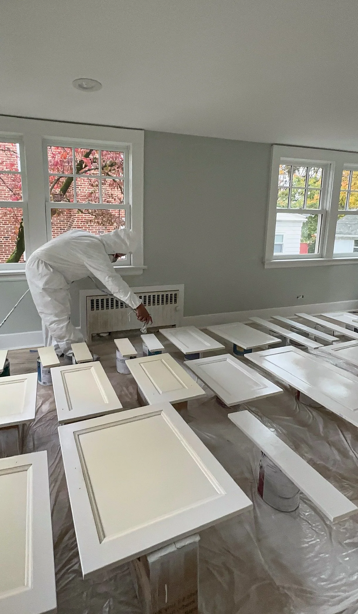 Person in protective suit painting kitchen cabinet doors white, placed on tables covered with plastic sheeting, in a room with large windows and trees outside.