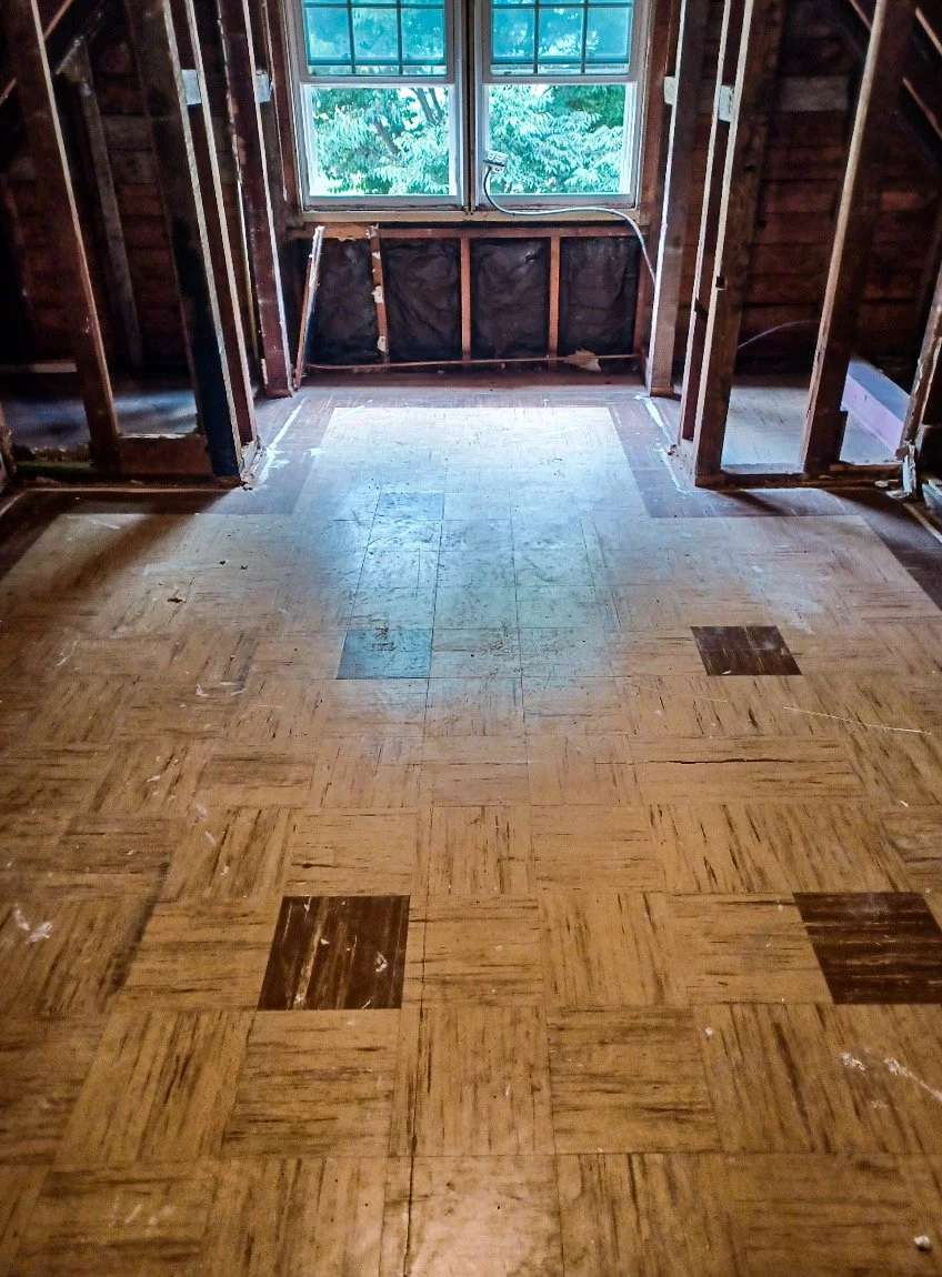 A room under renovation with exposed wooden framing, a window showing greenery outside, and a worn parquet floor.