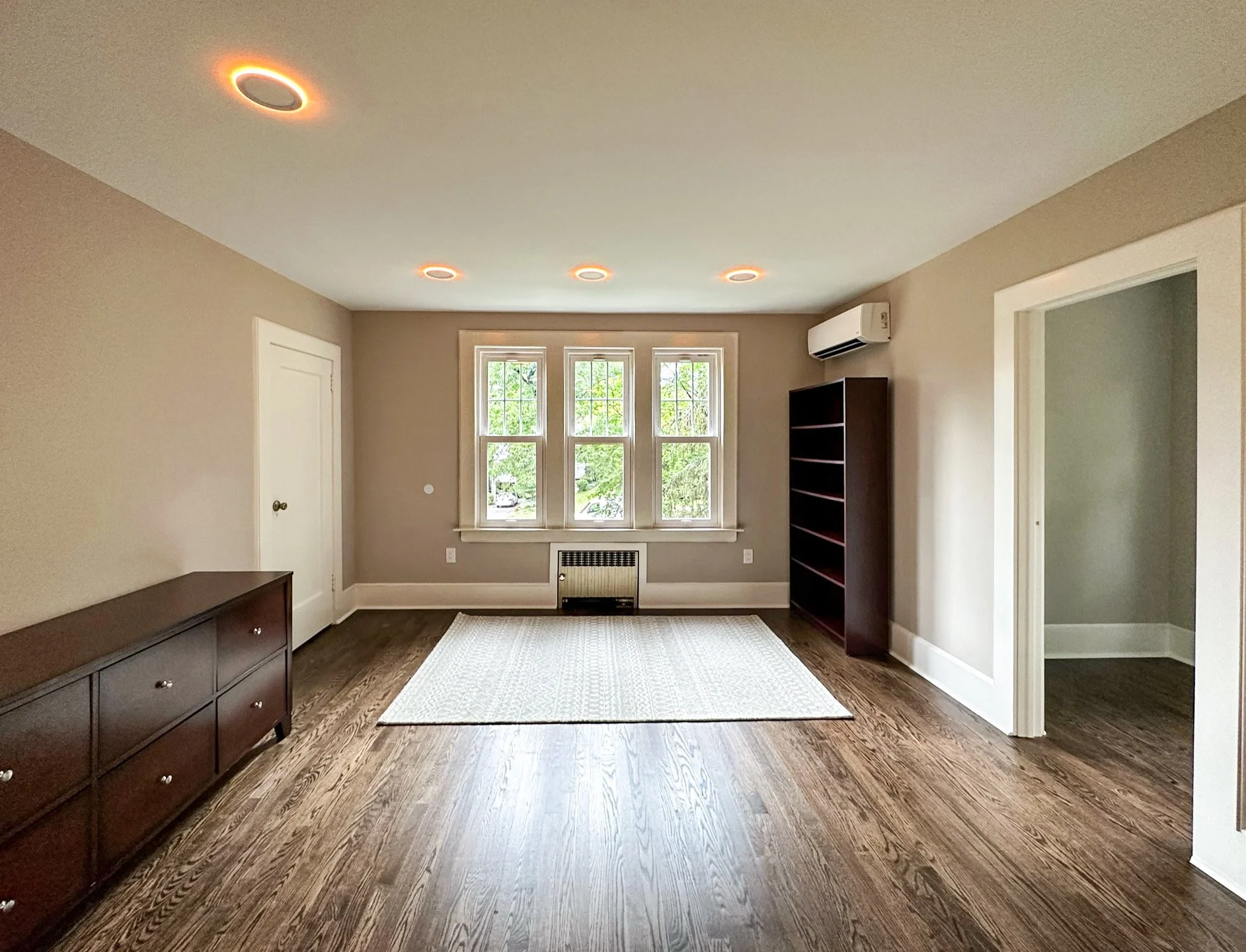 Empty living room with hardwood floors, beige walls, a large window, a white ceiling with recessed lighting, a dark wooden sideboard, a dark bookshelf, an air conditioning unit, a carpet in the center, and an open doorway to another room.