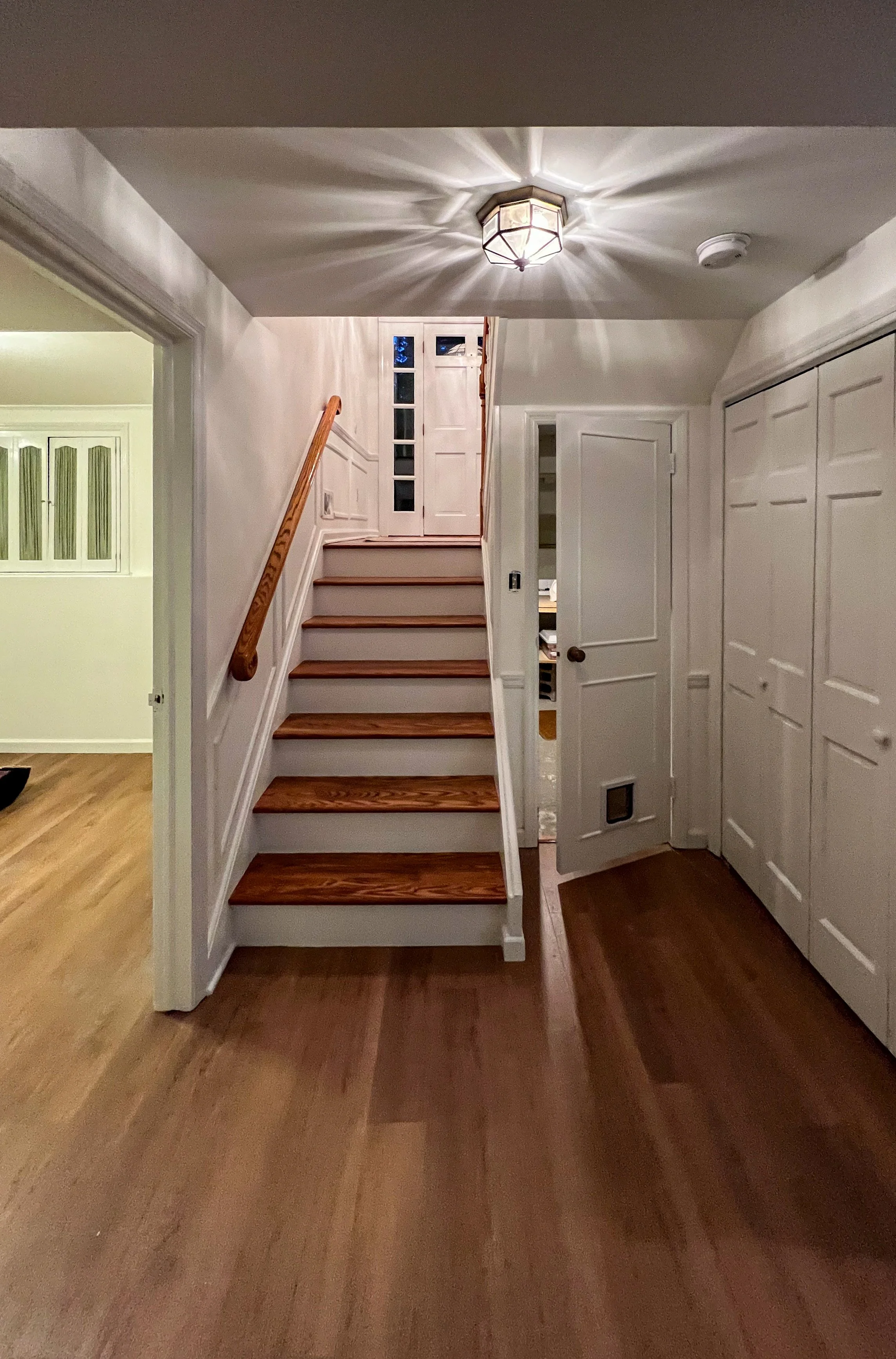 Interior view of a house showing a staircase with wooden steps and a handrail, a white door partially open, and built-in white cabinets on the right side, with a ceiling light casting shadows on the ceiling.