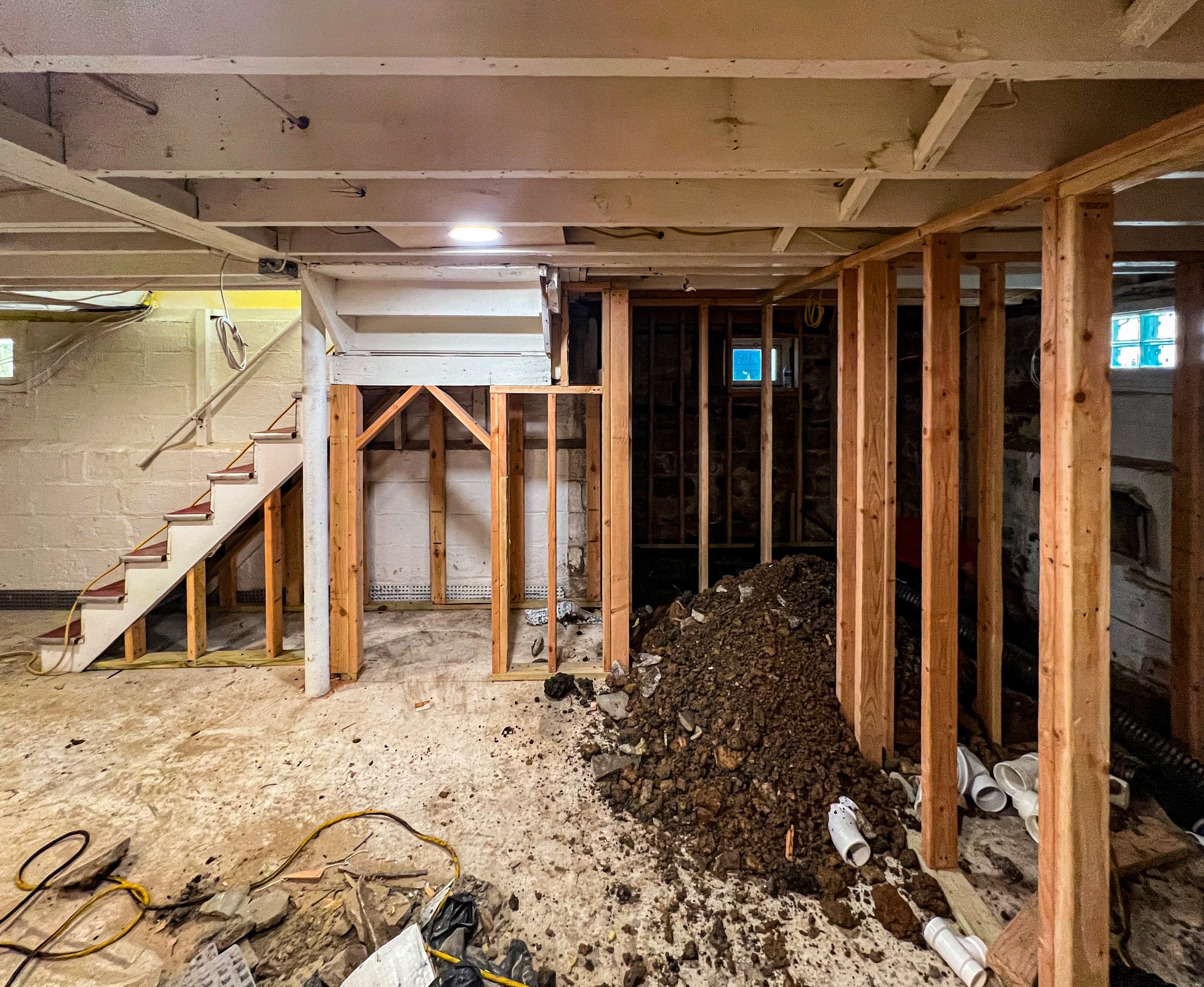 A basement under renovation with exposed wooden framing, a staircase, a pile of dirt and debris, and construction tools and materials scattered on the floor.