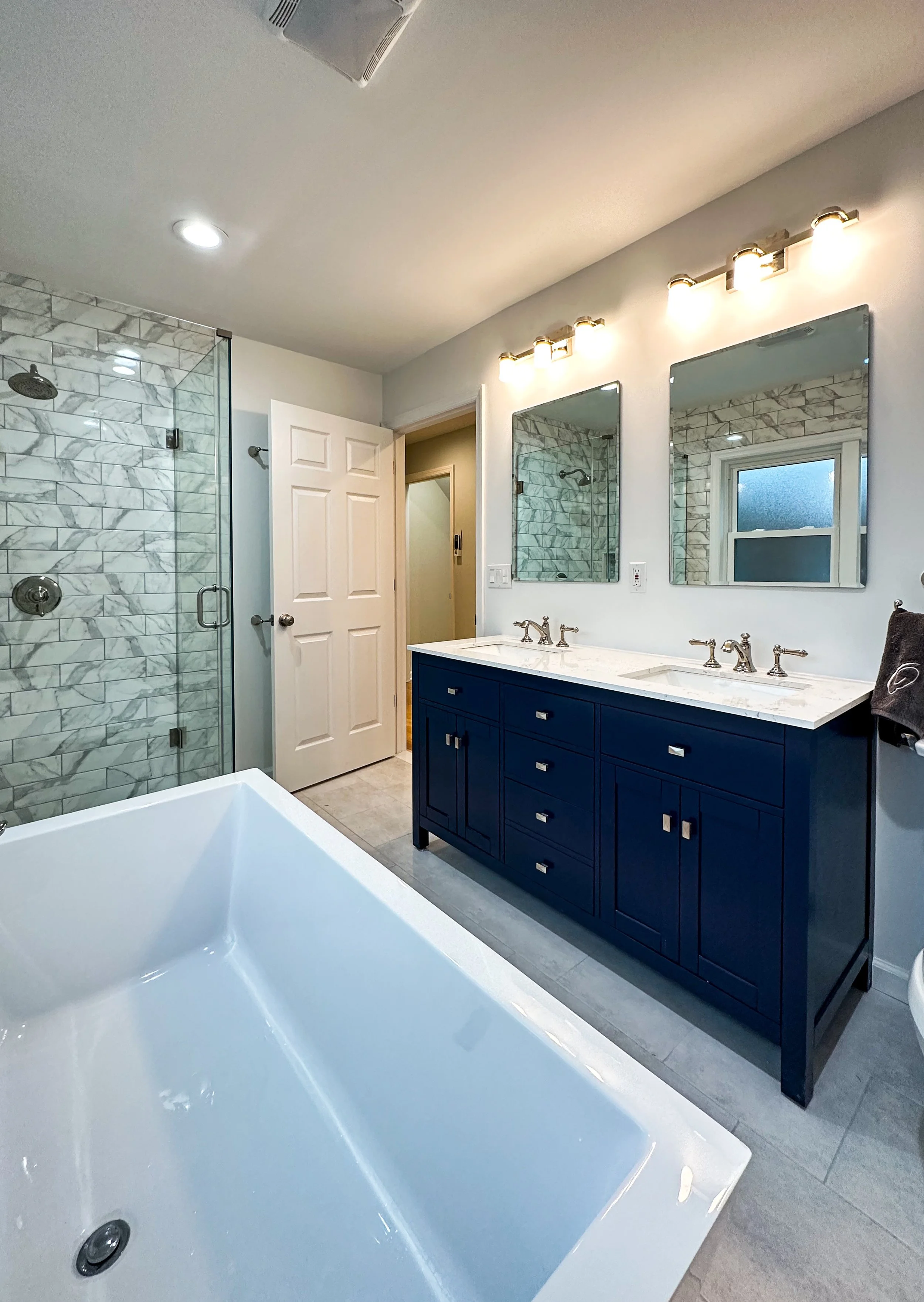 Modern bathroom with a large bathtub, a double vanity with two mirrors and lighting, a glass-enclosed marble shower, and a small window.