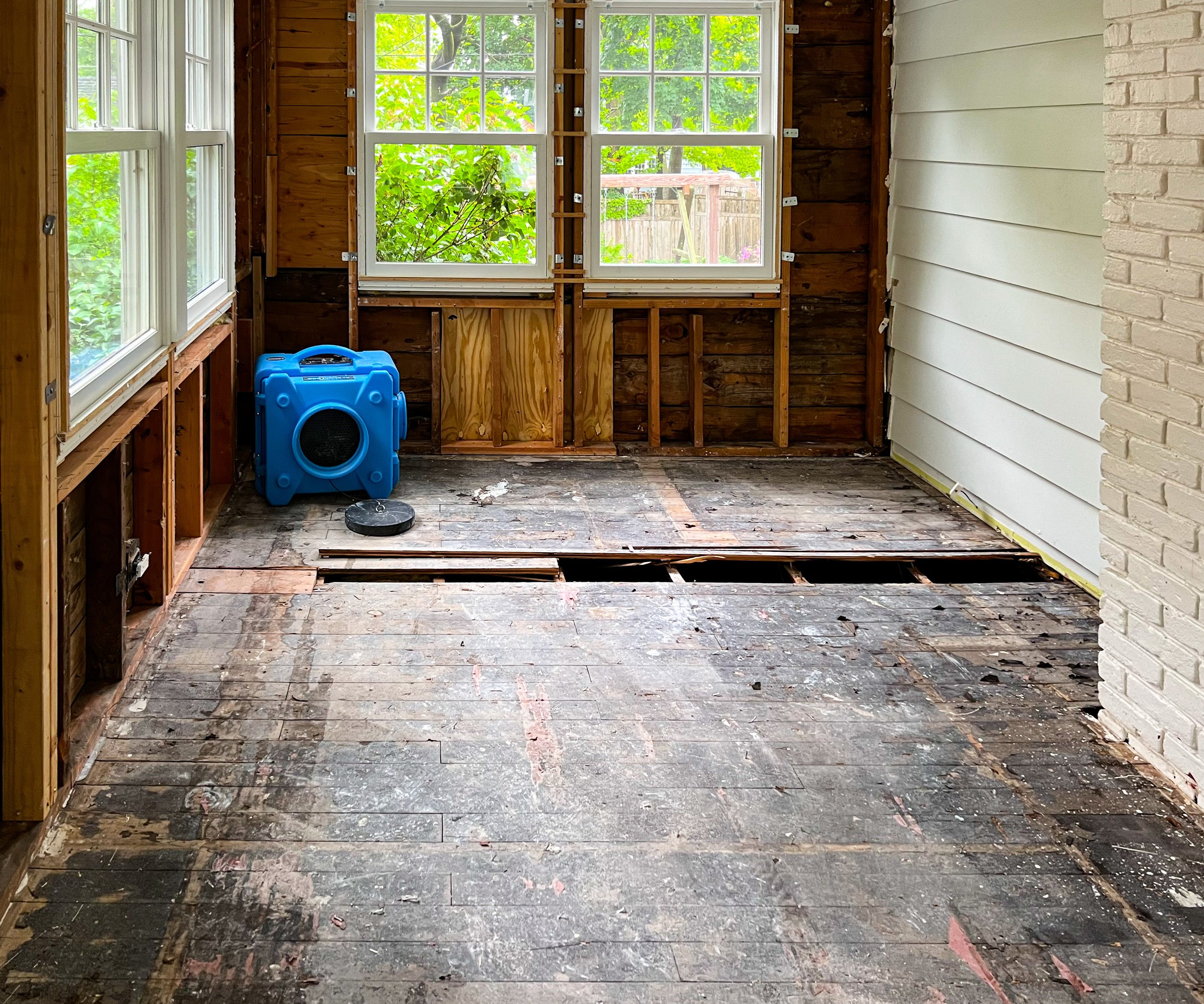 An unfinished room with exposed wooden flooring and walls, two windows overlooking a backyard with trees, and a blue speaker or portable stereo on the left side.