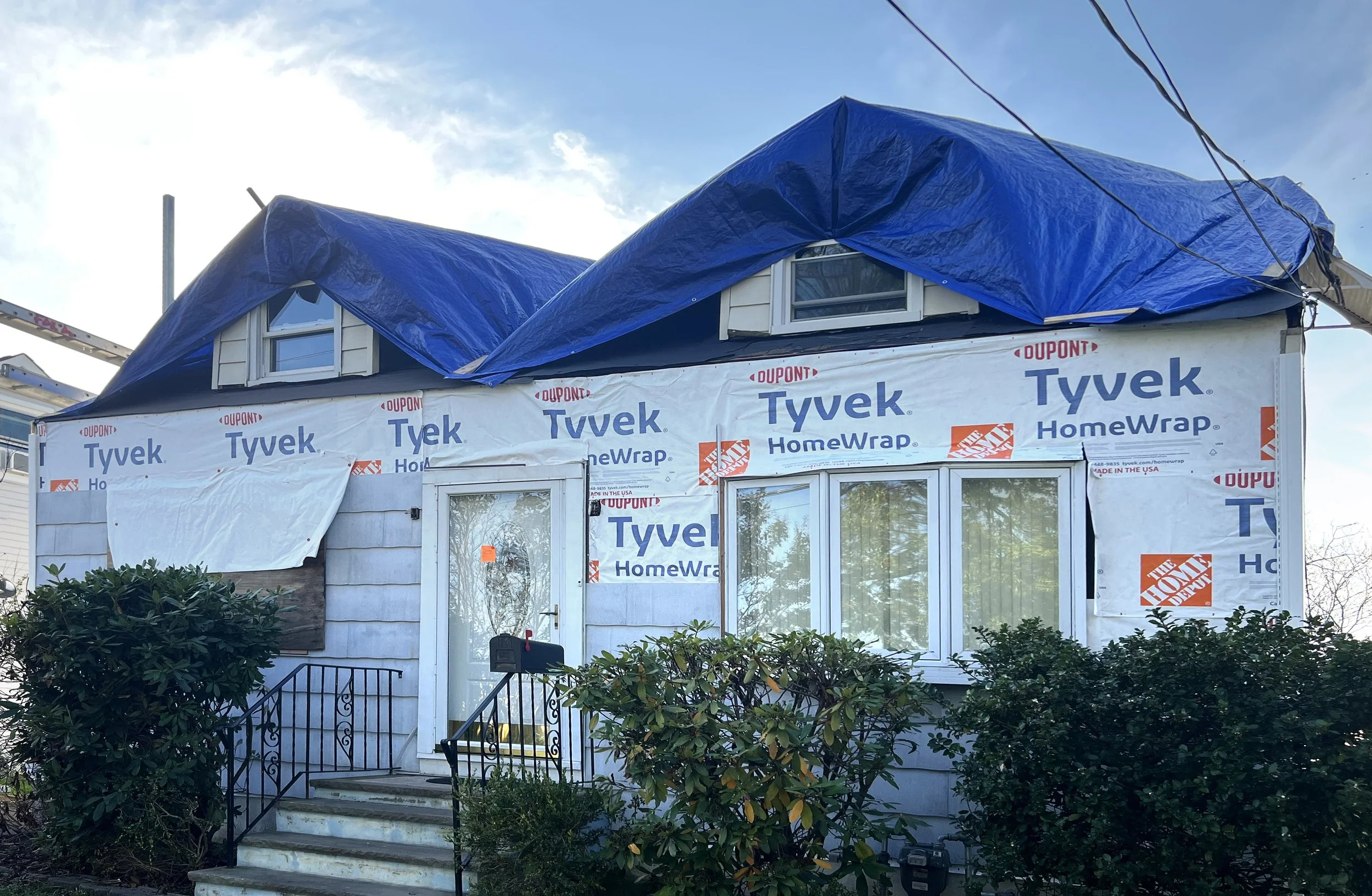 A house under construction with siding wrapped in Tyvek security wrap. Blue tarps cover the upper part of the roof, and the house has a front door with a mailbox and stairs leading up to it, surrounded by bushes.