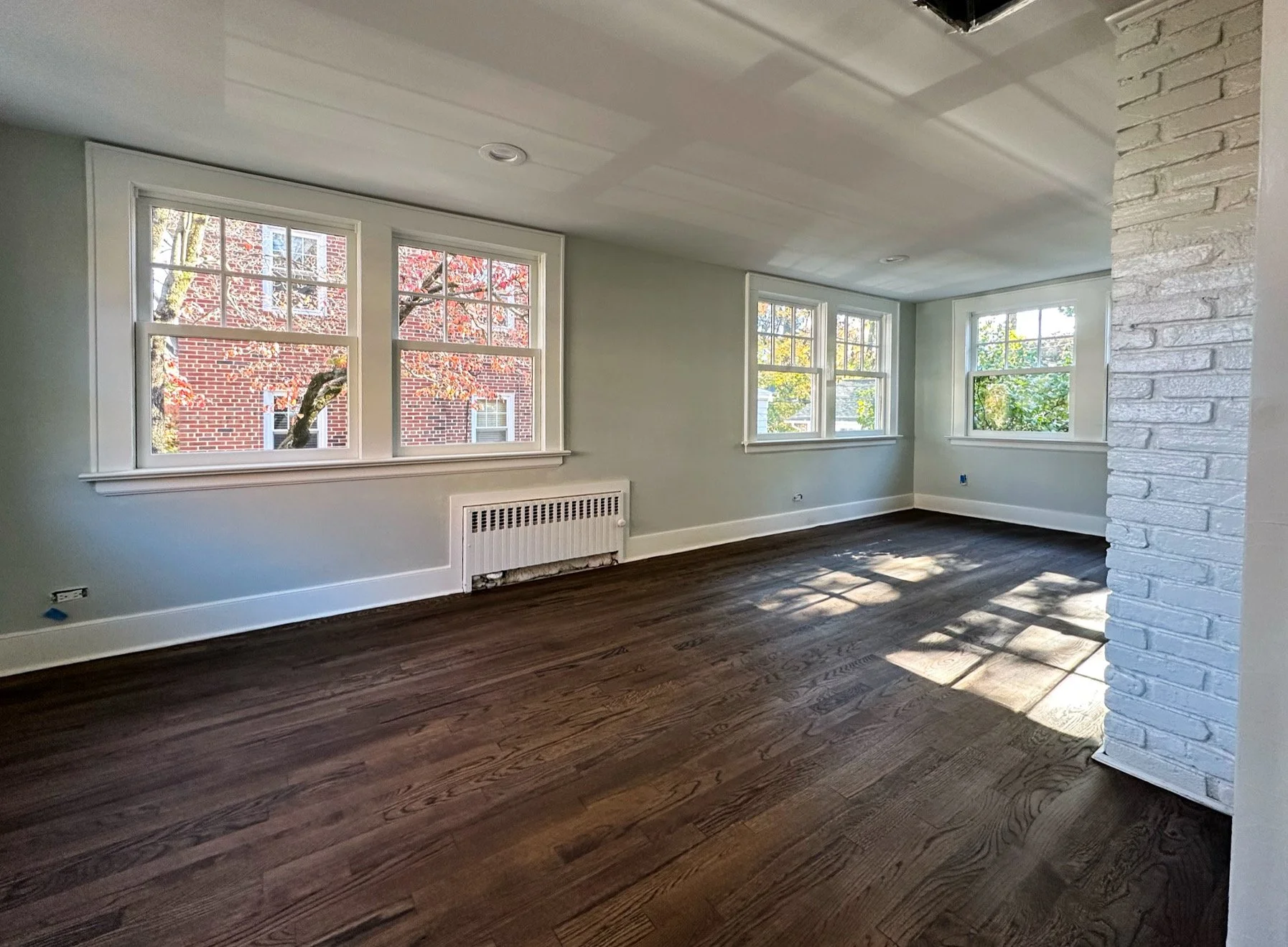 Empty living room with three large windows, white walls, dark hardwood floors, and a white brick column on the right, sunlight streaming through the windows.