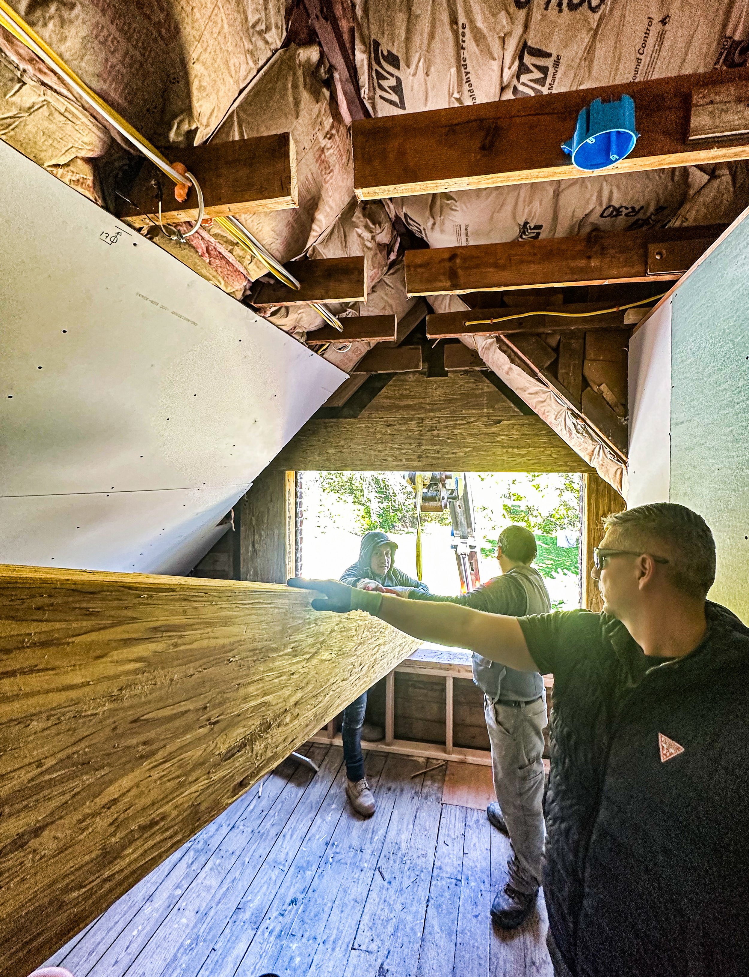 Construction workers installing a large wooden beam inside a room with unfinished walls and insulation, near a window with sunlight.