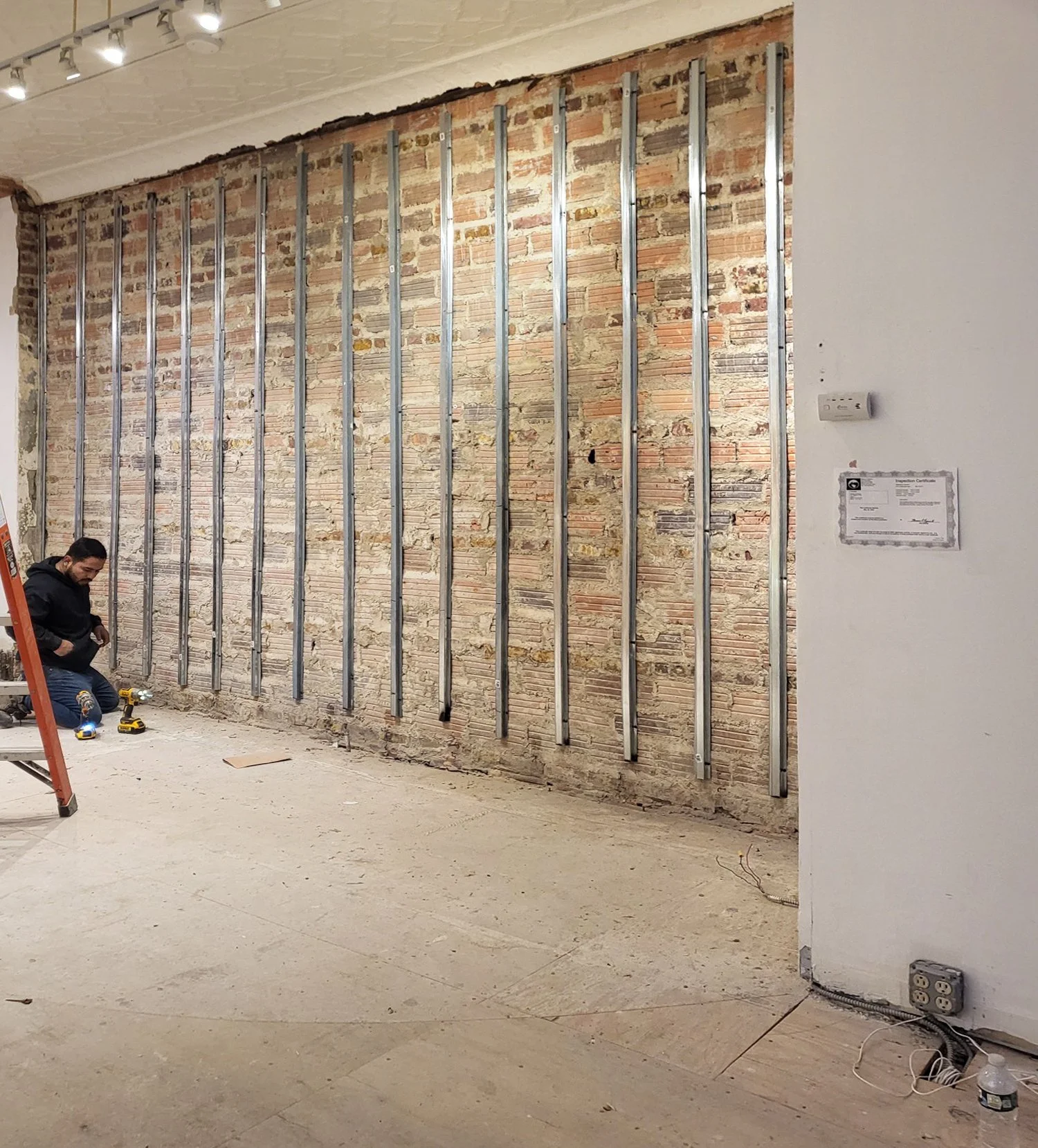 Interior of a room under construction with exposed brick wall and vertical metal studs. Two workers are on the floor, one sitting near a ladder with tools around.