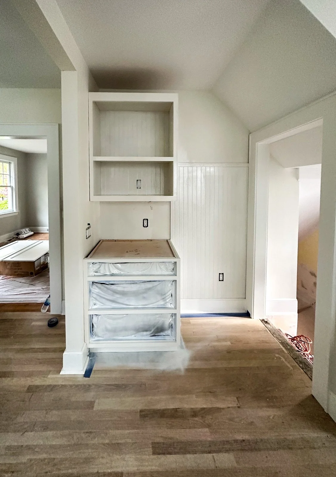 Interior of a house under renovation, showing a built-in white cabinet with open shelves and drawers covered with protective plastic, light-colored wood flooring, and white walls with beadboard paneling.