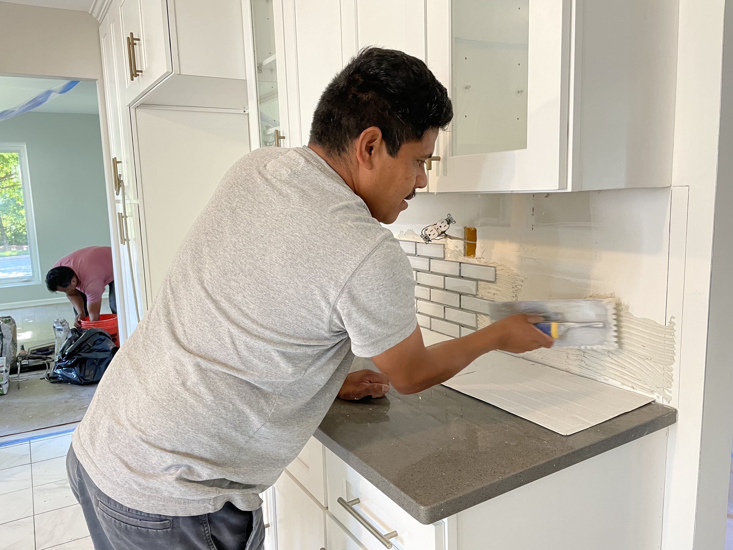 A man applies white tile adhesive to a kitchen wall with notched trowel, preparing to install tiles, with a woman working in the background near a window.