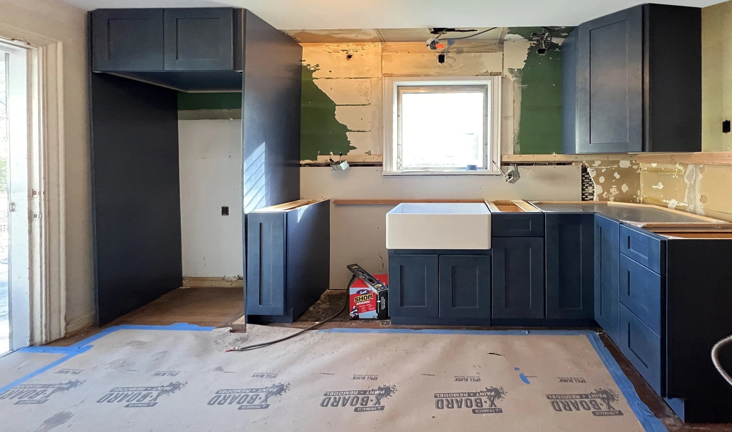 Kitchen under renovation with dark blue cabinetry, a farmhouse sink under a window, and unfinished walls with exposed drywall and paint patches.