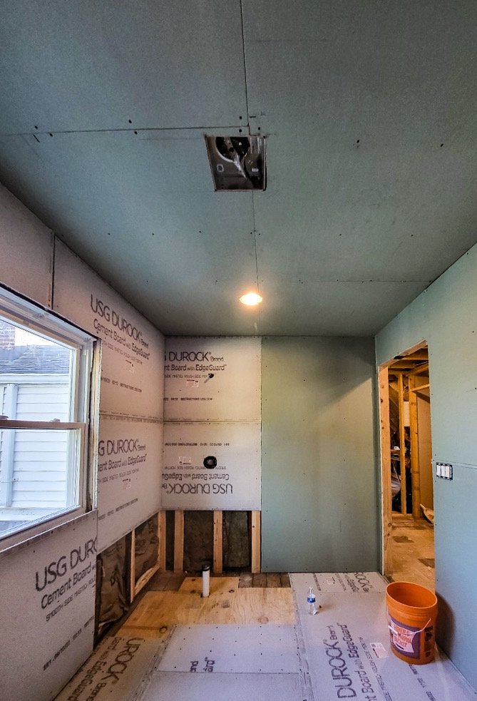 Interior room under construction with drywall and wooden framing, partially finished walls, a window on the left, a ceiling light, and construction materials including an orange bucket and a water bottle on the floor.