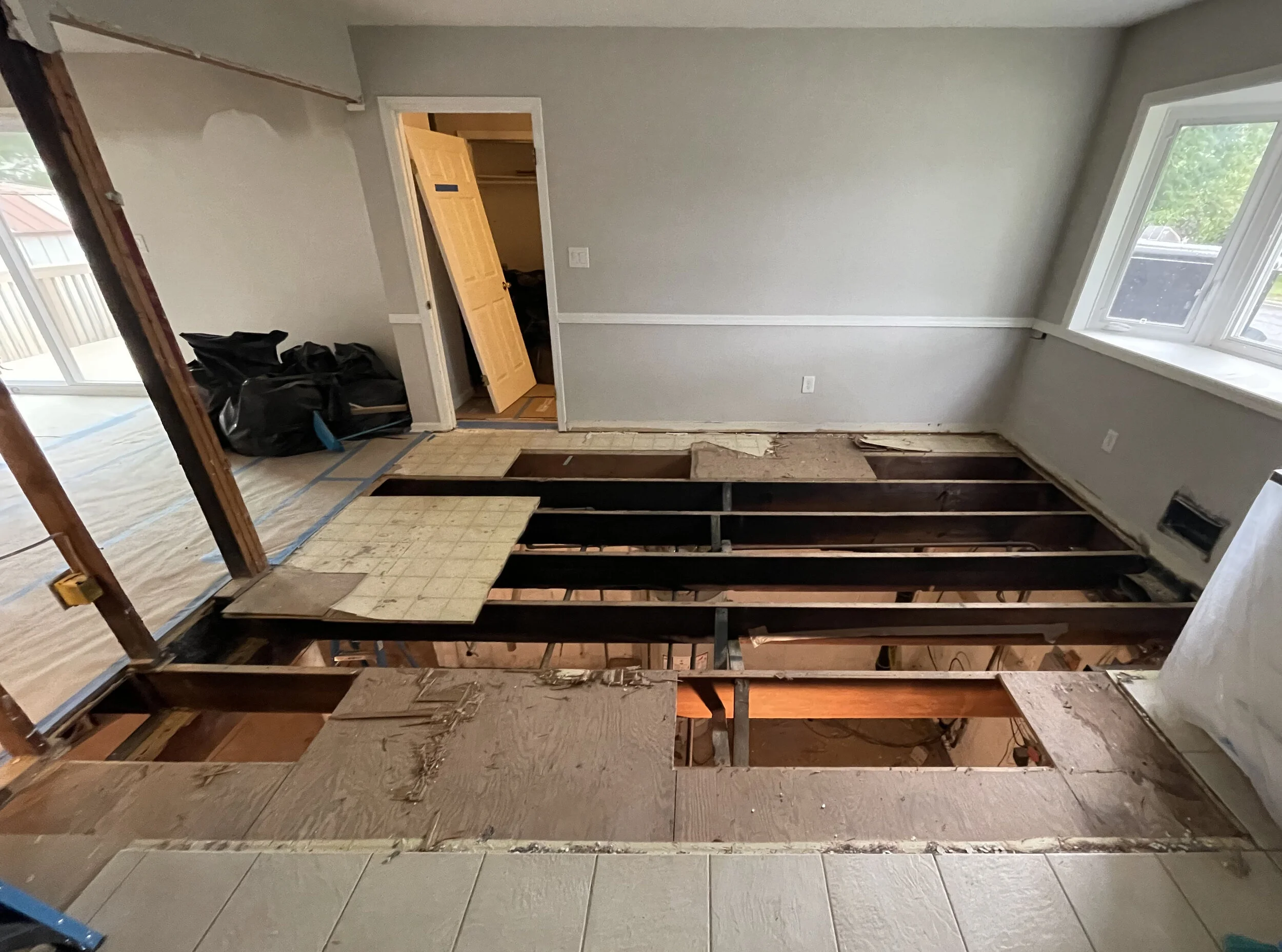 Room under construction with the floor removed, exposing the wooden beams and subfloor. The room has a large window, a door leading to a closet, and black garbage bags in the corner.