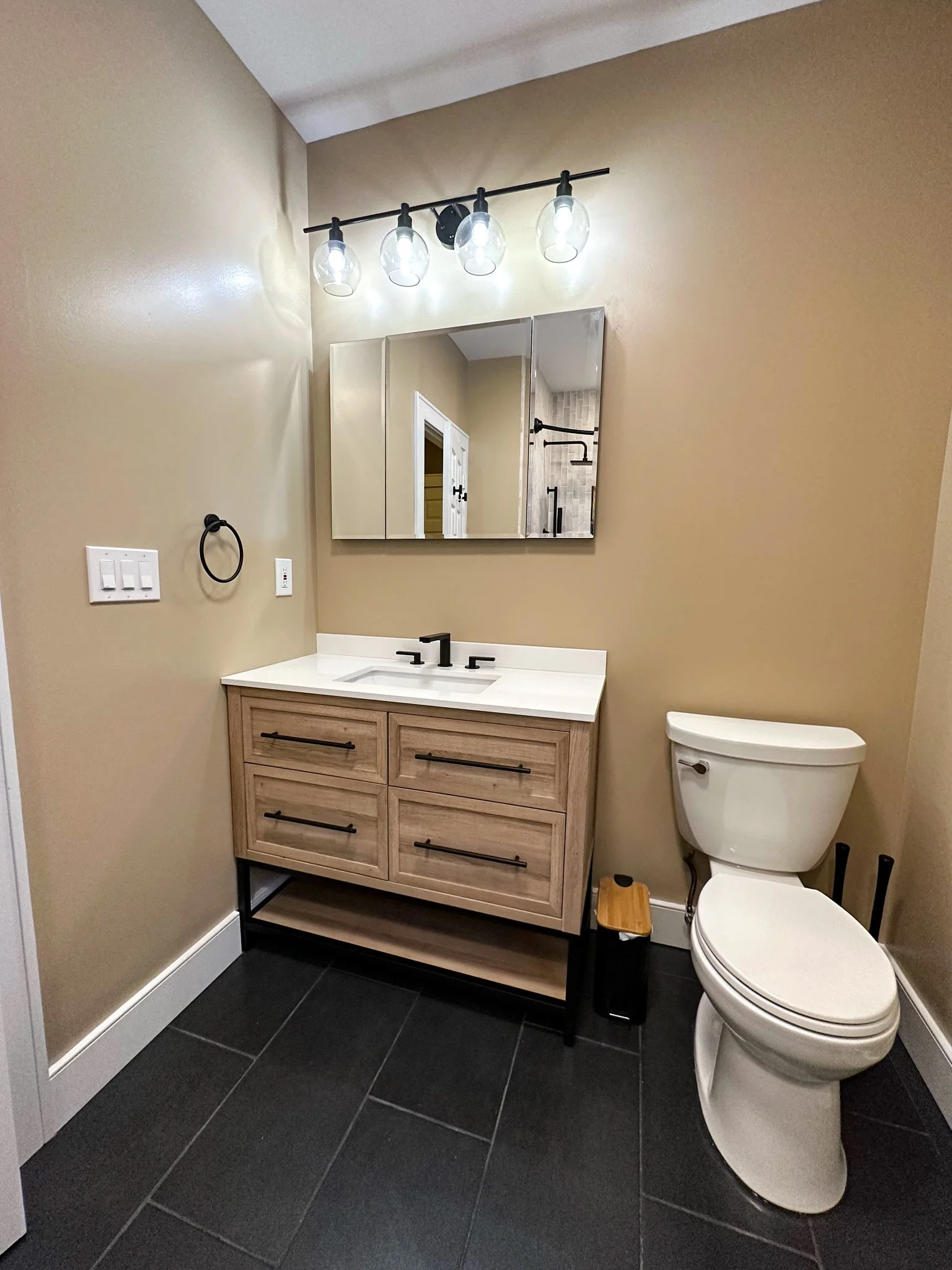 Modern bathroom with beige walls, black tile flooring, wooden vanity with black handles, white countertop, bowl sink, black faucet, large mirror, four-light fixture, and toilet.